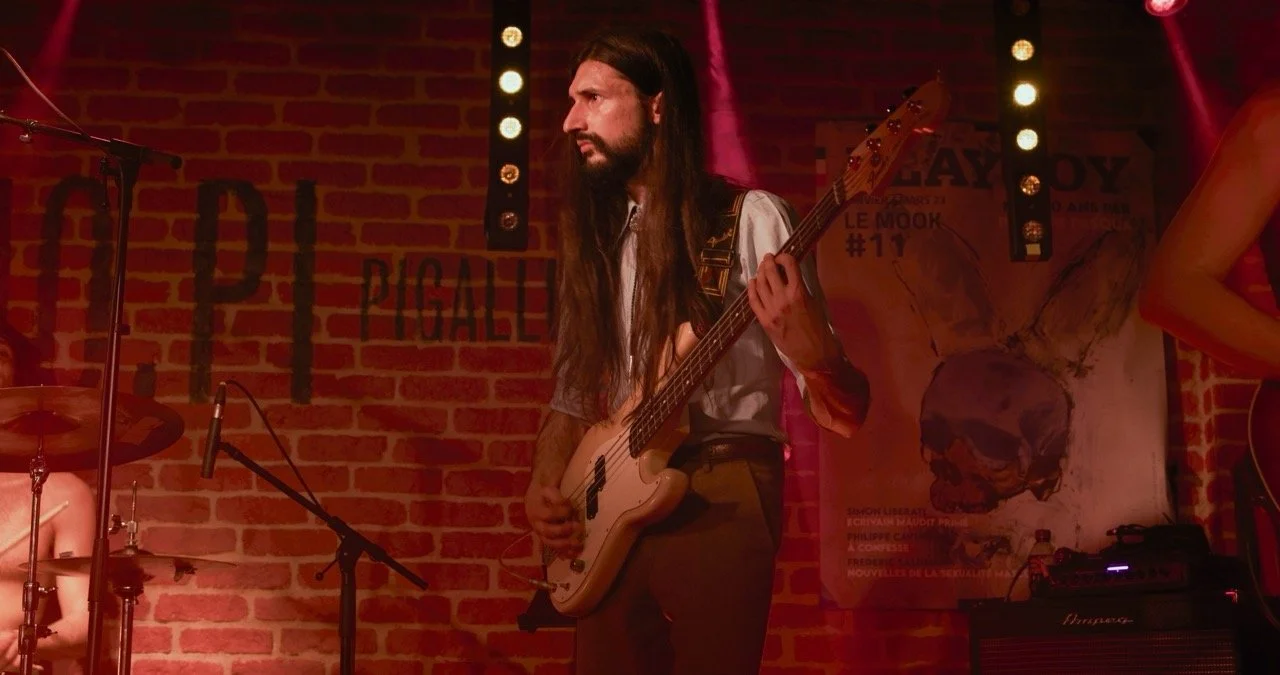 Un homme avec une guitare électrique sur scène dans un bar avec un mur en briques en arrière-plan.