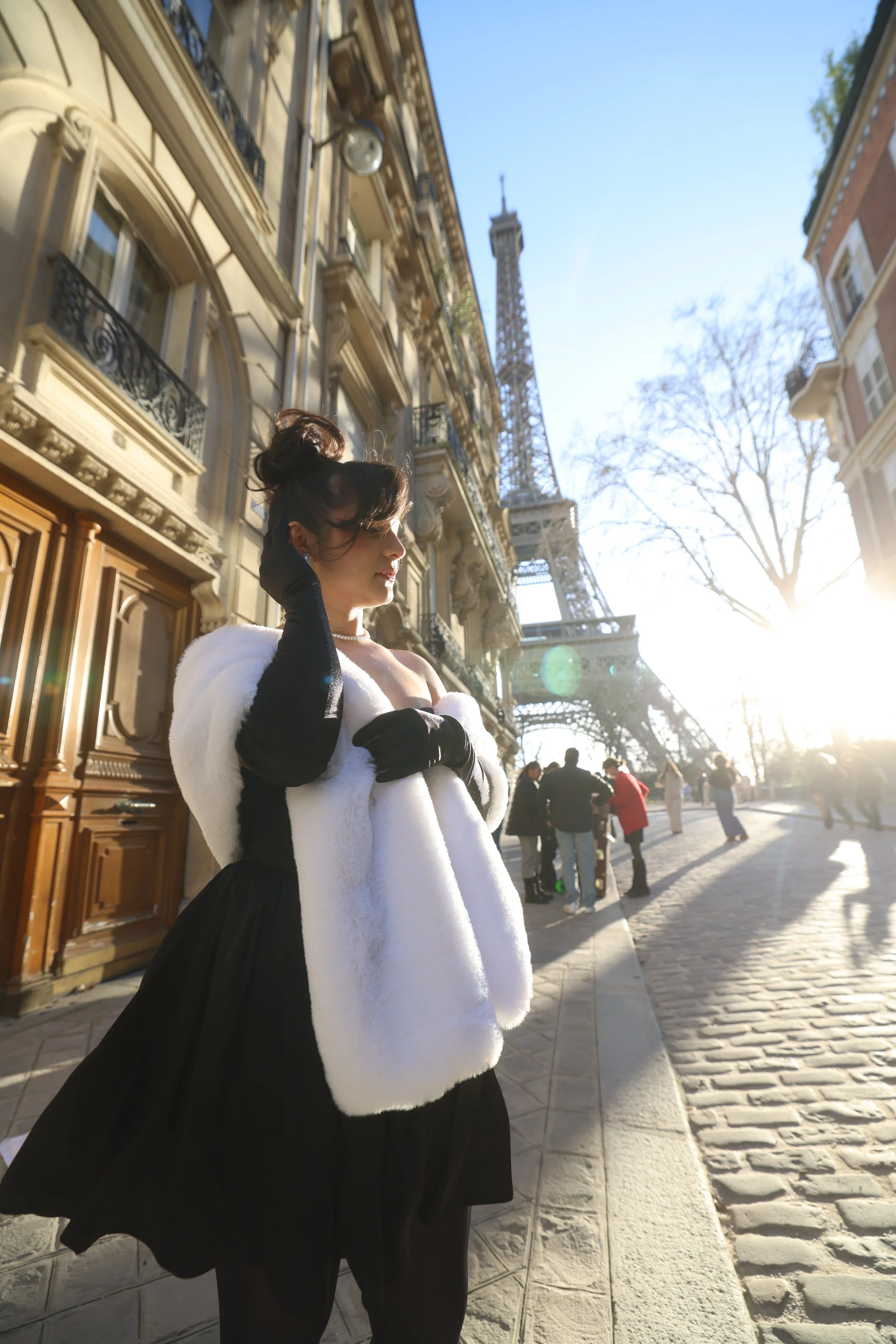 Une femme habillée élégamment, portant une robe noire, une étole blanche en fourrure et des gants noirs, se tient sur un trottoir parisien avec la tour Eiffel en arrière-plan, en pleine journée ensoleillée.