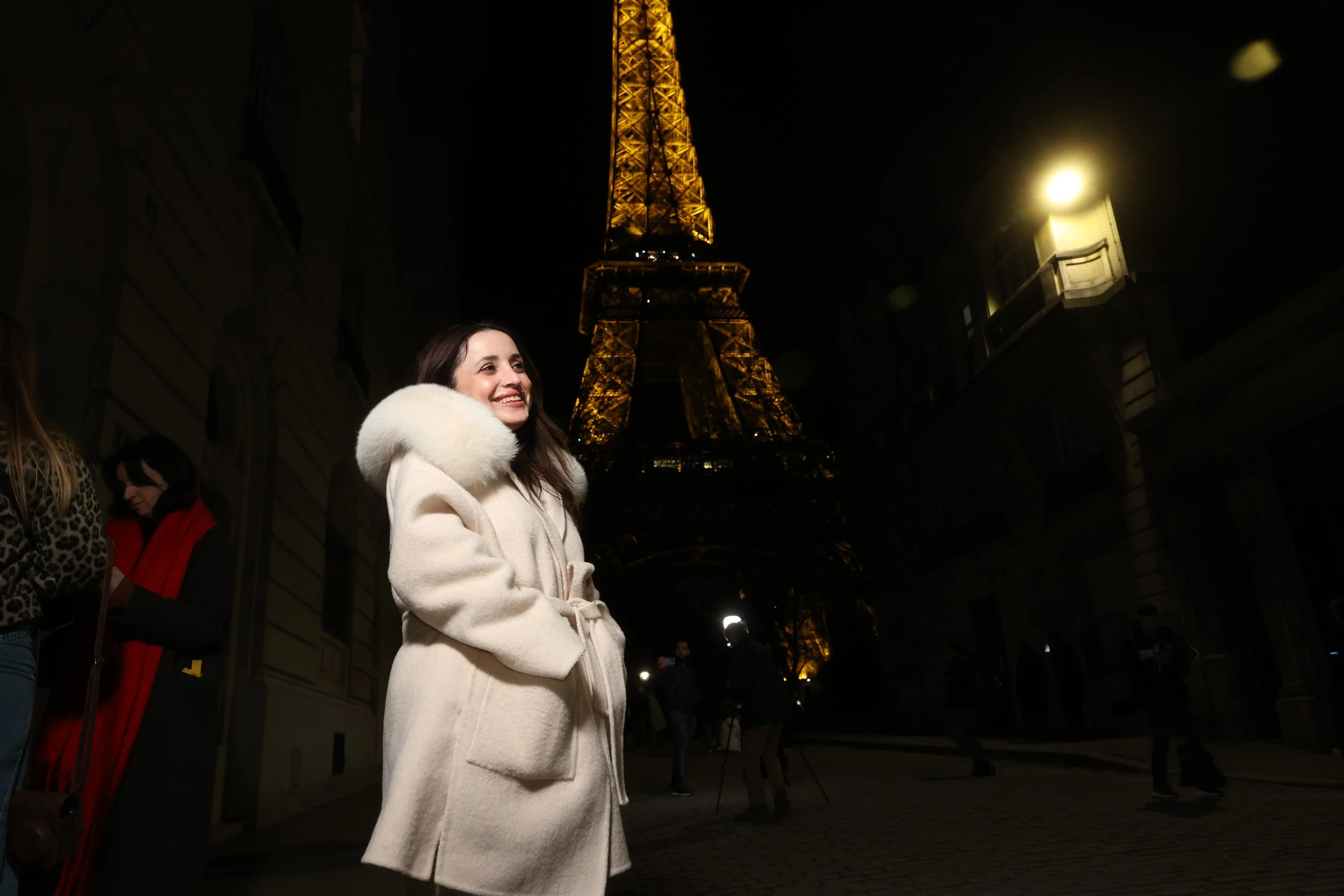 Femme souriante portant un manteau blanc près de la Tour Eiffel illuminée la nuit à Paris.