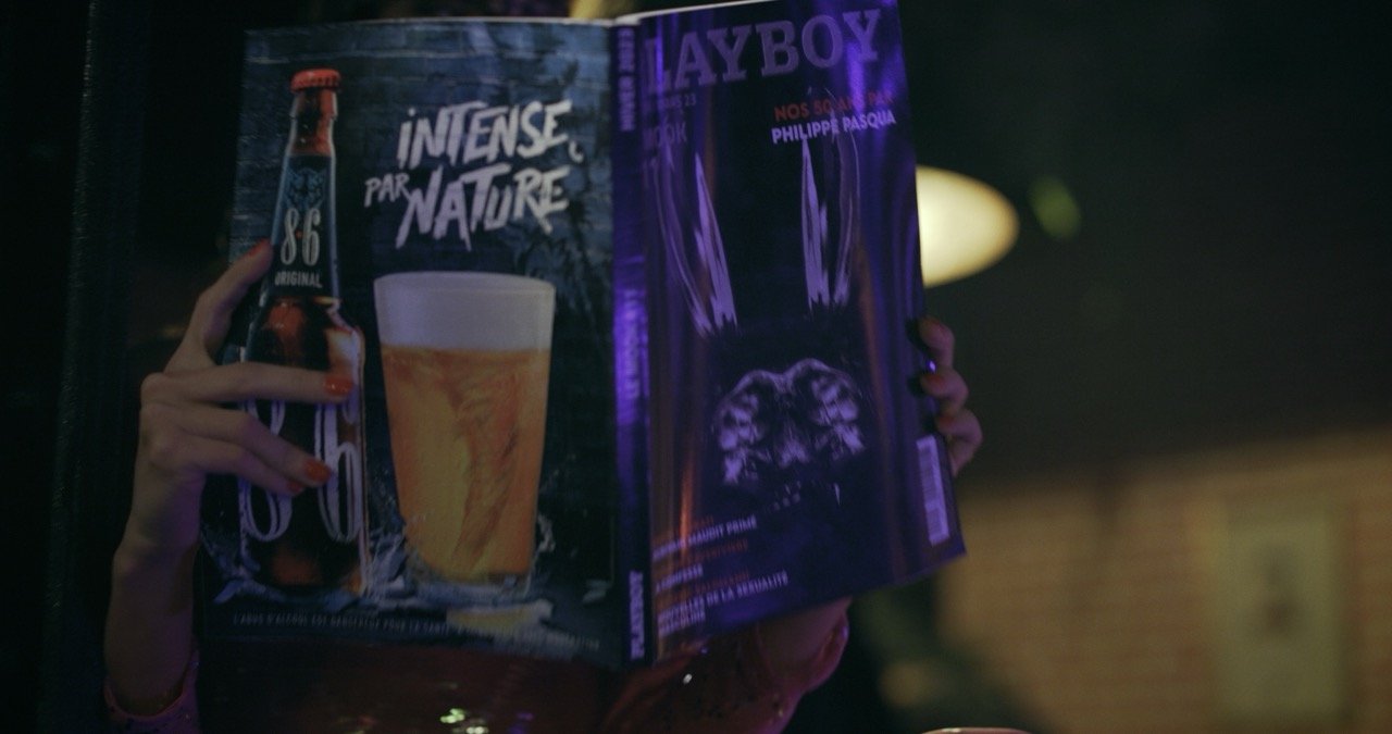 Une personne tient une affiche ou un magazine ouvert avec une publicité pour une bière, un verre de bière blonde, et un magazine Playboy avec une couverture violette.