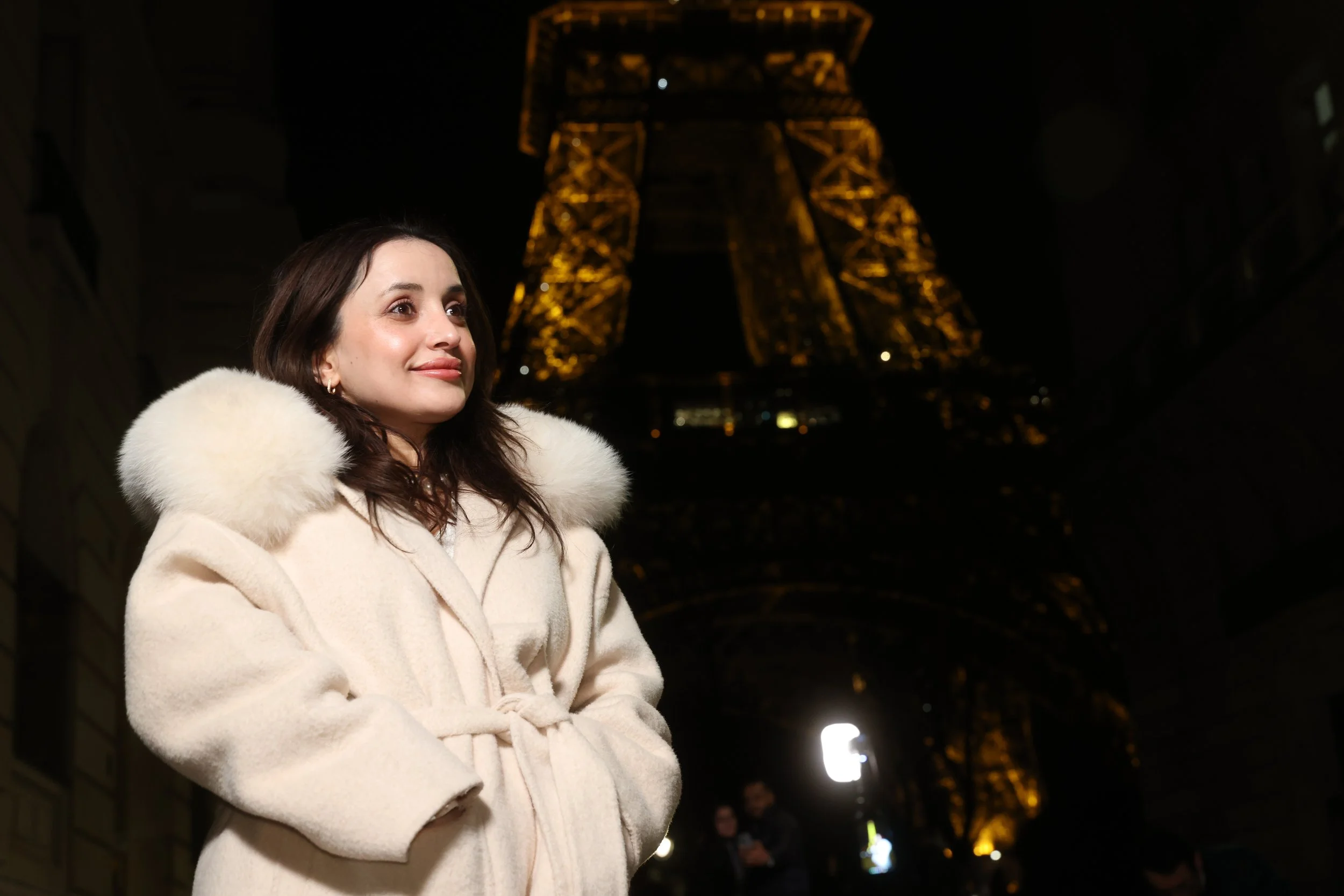 Une femme portant un manteau blanc avec une capuche en fourrure, se tenant devant la Tour Eiffel illuminée la nuit à Paris.