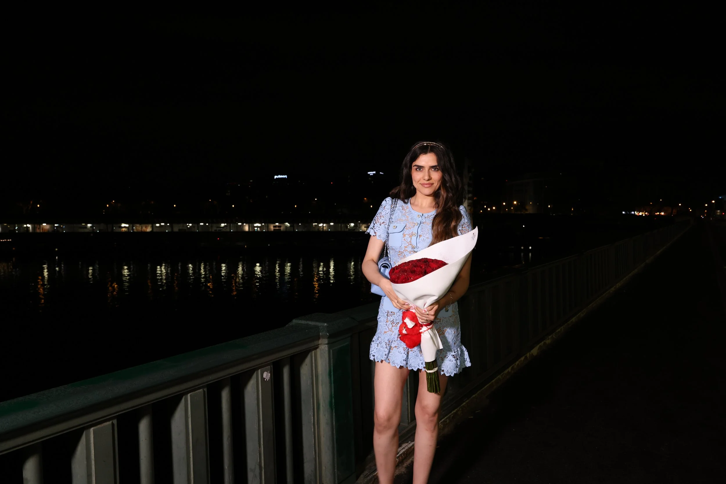 Jeune femme avec une robe bleue en dentelle, tenant un bouquet de fleurs rouges, debout sur un pont la nuit avec des lumières derrière elle.