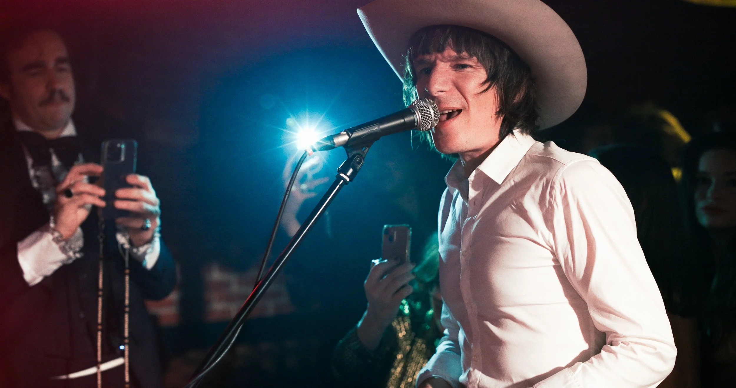 Un chanteur portant un grand chapeau blanc chante dans un micro lors d'une performance en live, entouré de quelques spectateurs qui prennent des photos avec leurs téléphones.