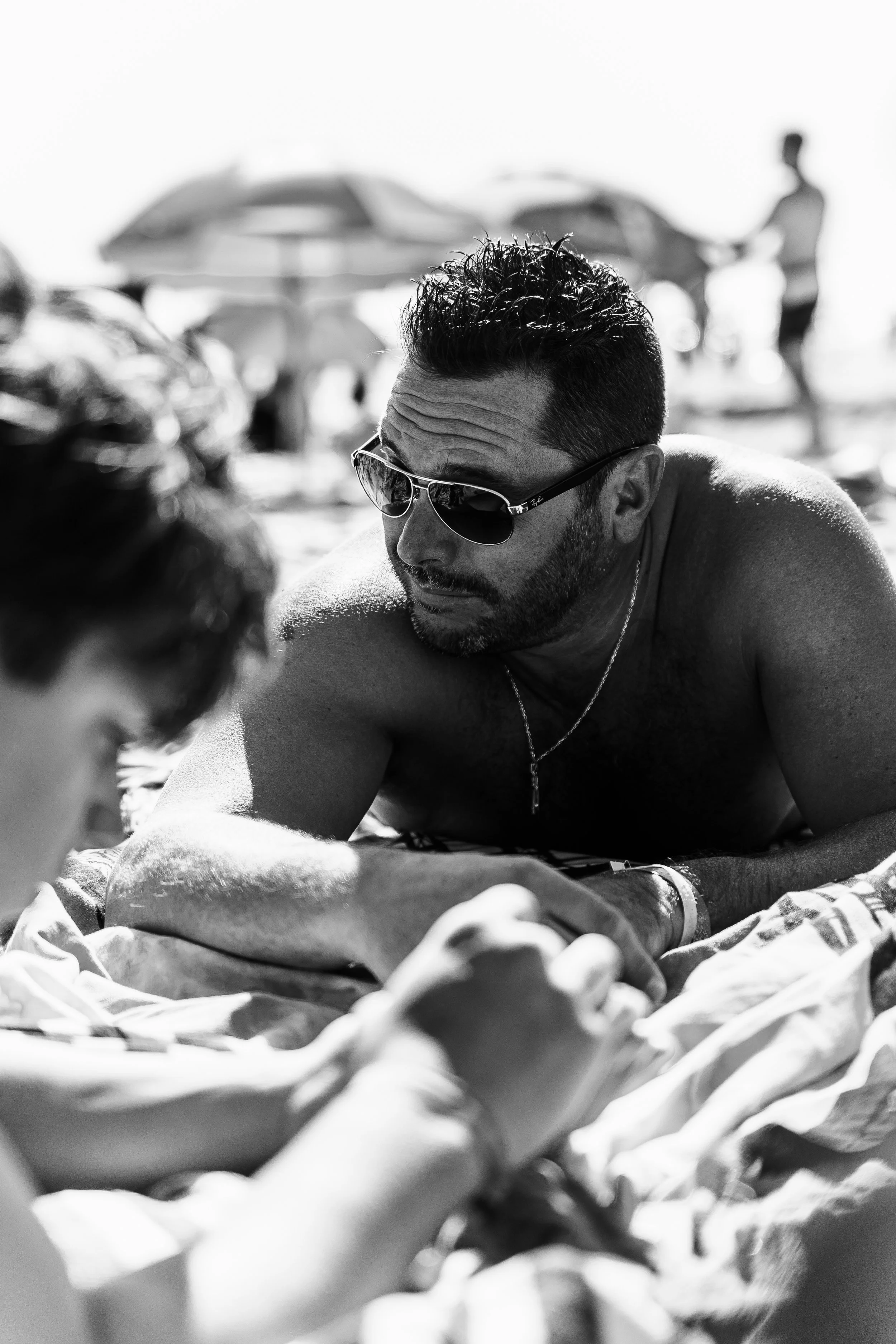 Un homme surfer avec des lunettes de soleil, allongé sur la plage, en train de jouer à un jeu avec un enfant, avec d'autres personnes en arrière-plan sous des parasols.