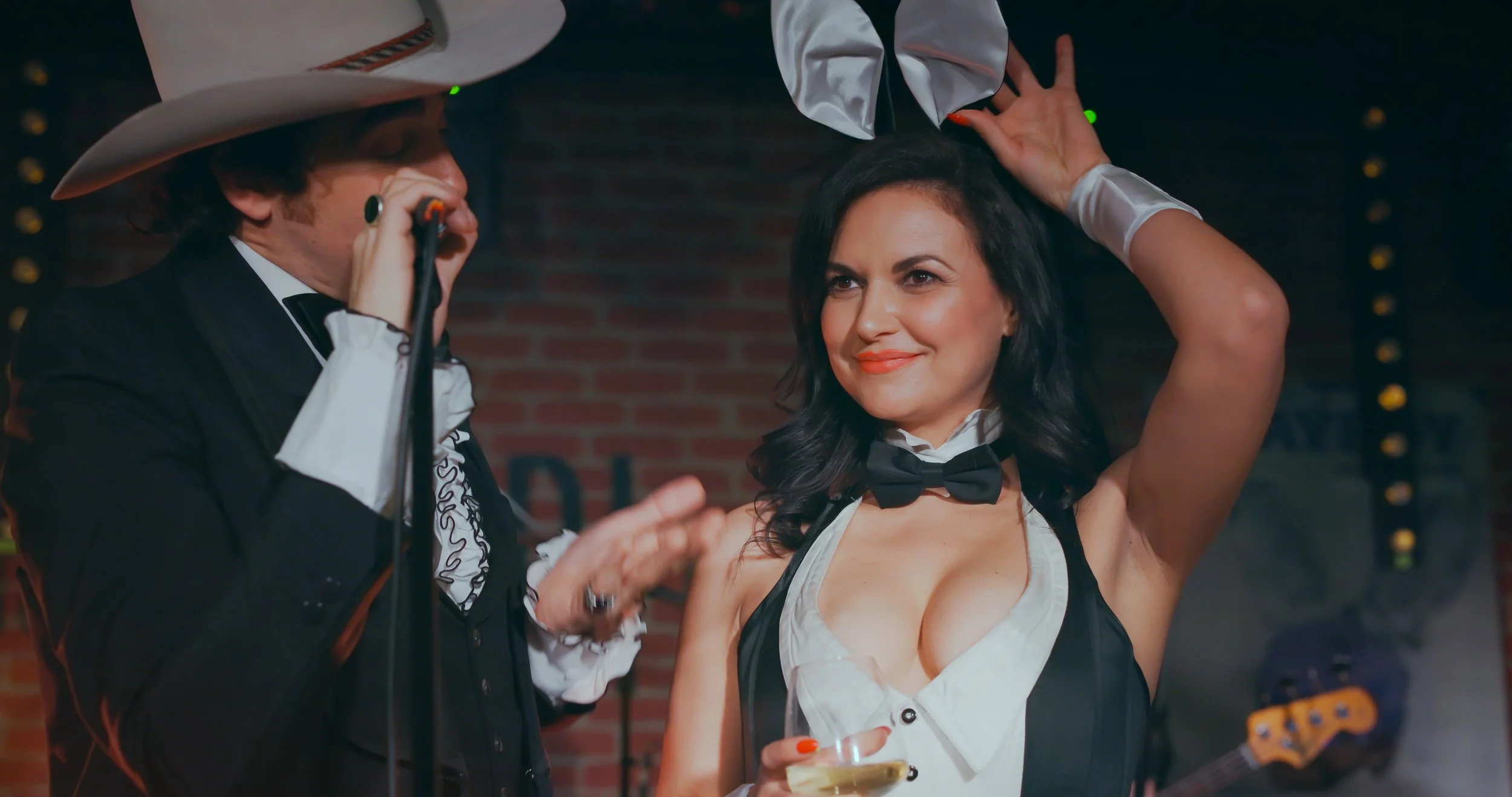 Une femme en tenue de barman ou serveuse habillée de manière séduisante avec un nœud papillon noir, qui sourit en tenant un verre de vin blanc lors d'une fête ou événement, avec un homme habillé comme un musicien ou chanteur avec un gros chapeau, dan