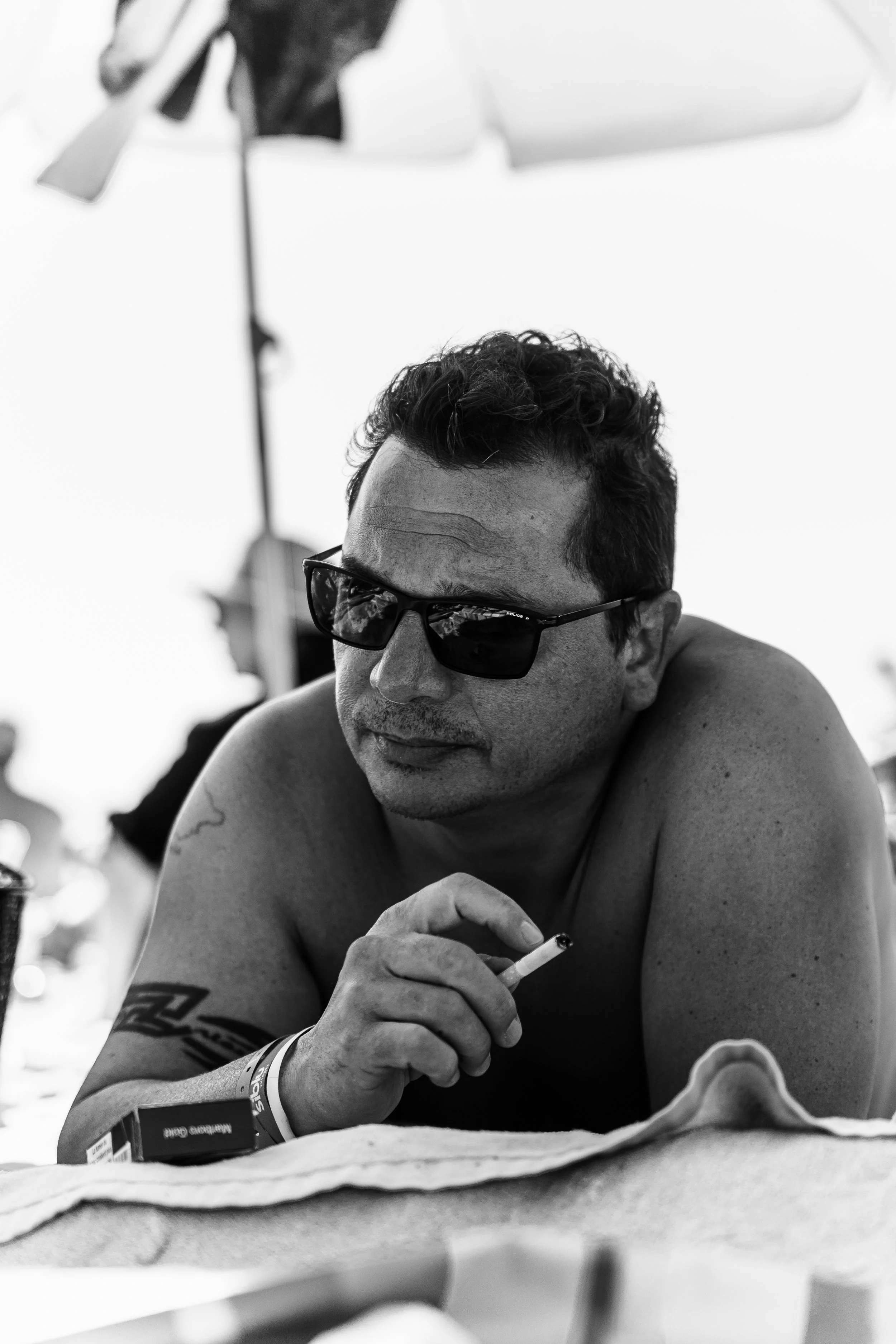 Homme portant des lunettes de soleil, assis, tenant une cigarette, avec un tatouage sur le bras, en arrière-plan une tente et un parasol, en noir et blanc.