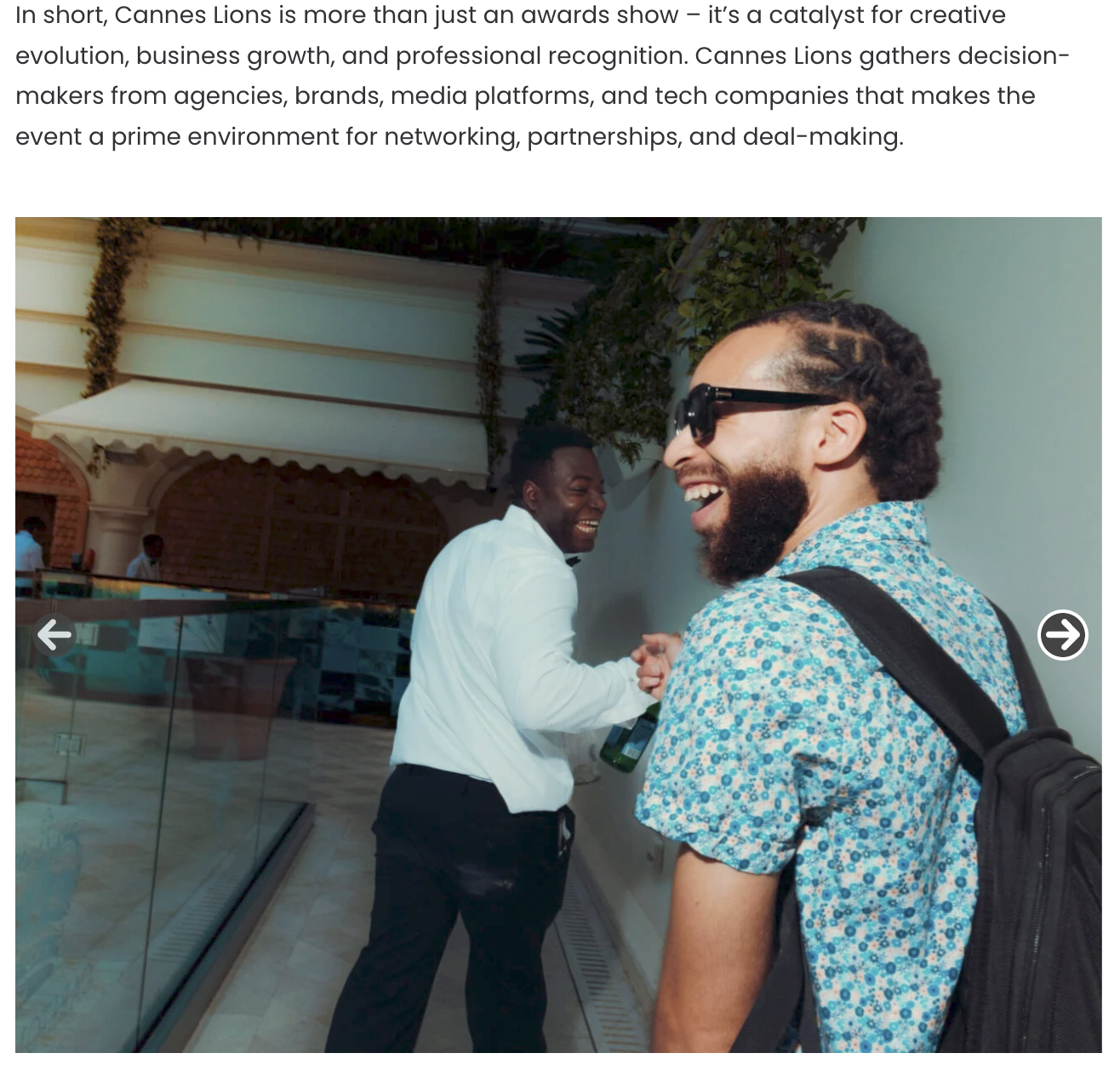 Deux hommes souriants en train de rire lors d'un événement, l'un avec une chemise à motifs bleus et lunettes de soleil, l'autre en chemise blanche, dans un cadre extérieur avec des plantes et une structure en fond.