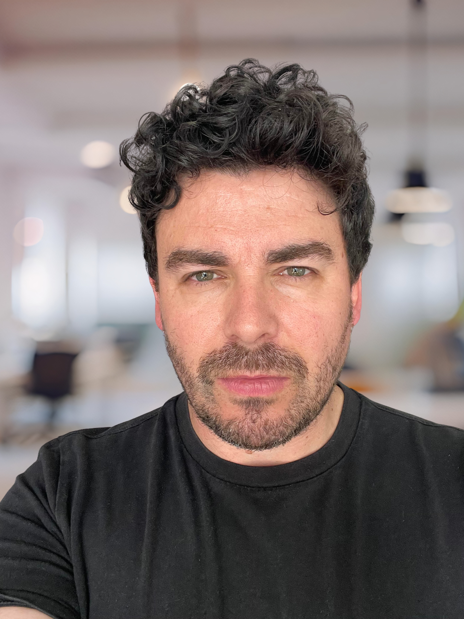 Close-up of a man with curly dark hair, light facial hair, wearing a black T-shirt, in a well-lit indoor office setting.
