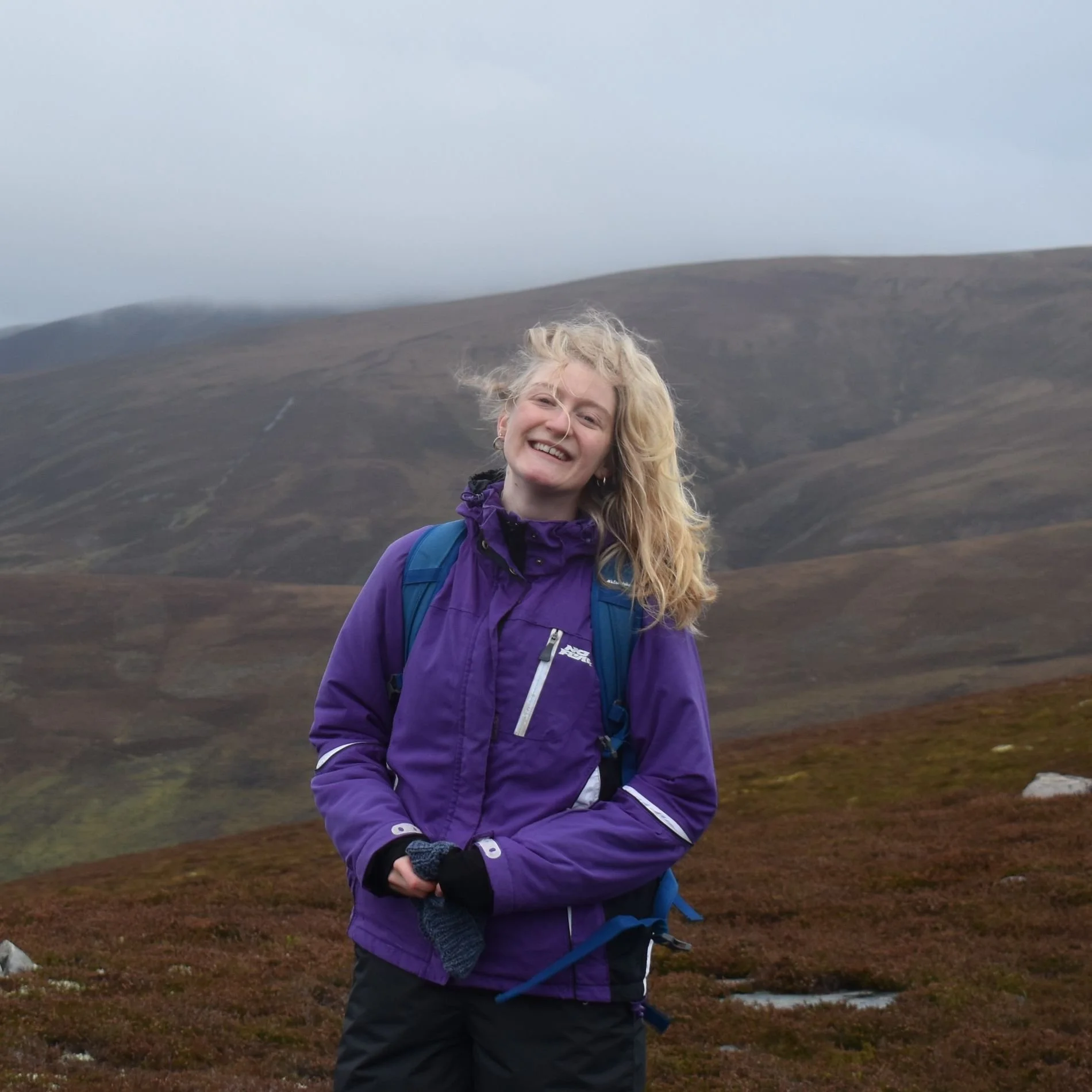 Photo of me - a young, blonde-haired woman in a purple coat - smiling at the camera and standing on a windswept moorland peak.