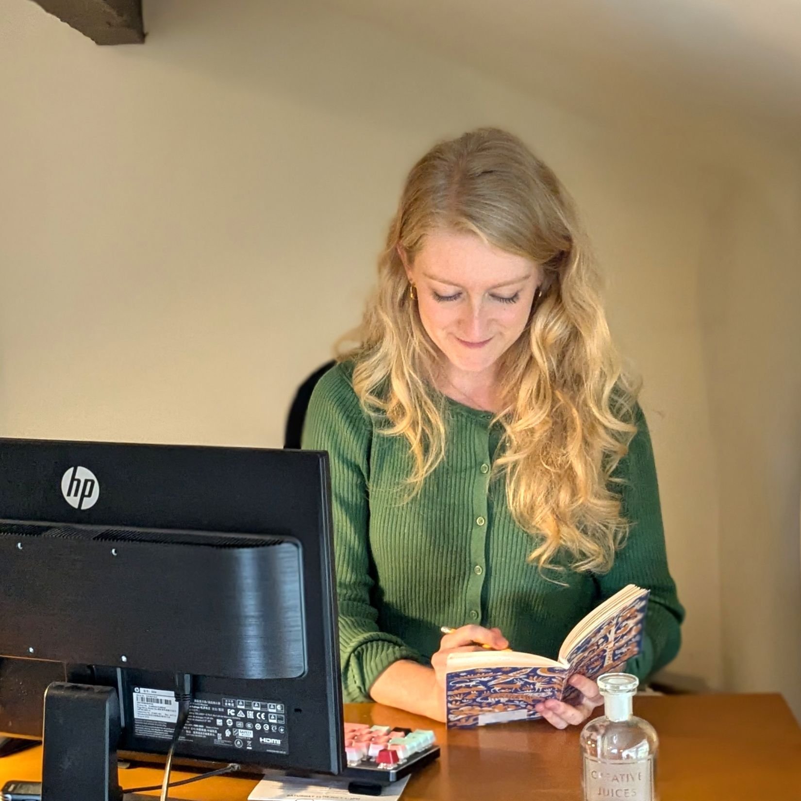 Photo of me - a young woman with blonde hair, wearing a green top, looking down at the notebook I'm writing in, while doing some freelance proofreading.