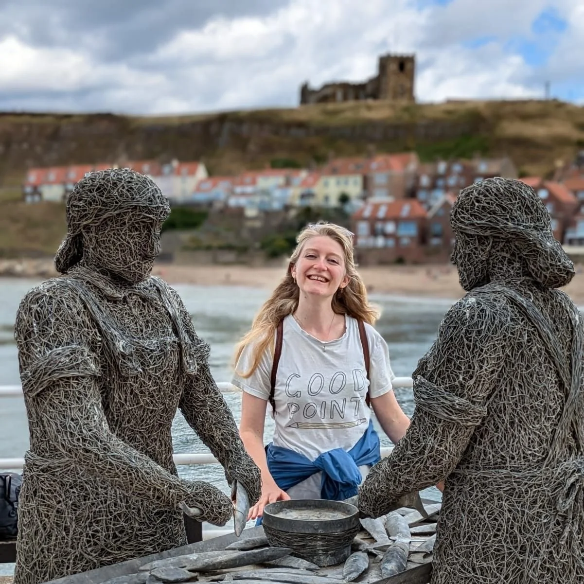 Photo of me - a smiling blonde-haired woman - standing by the fishwives statues on the Whitby Heritage Trail.
