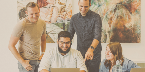Photo of four people smiling and looking at someone's laptop together in a professional setting.