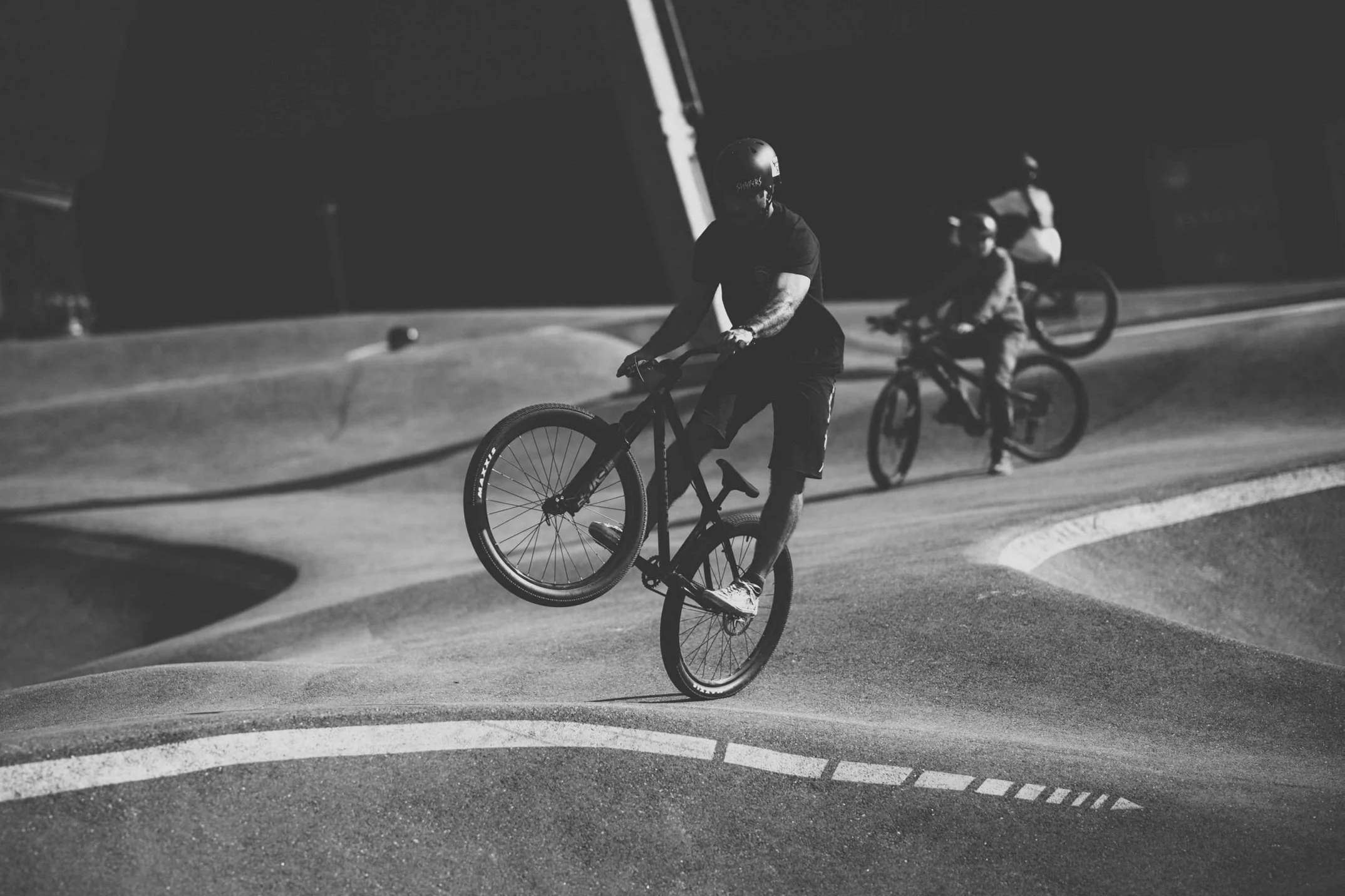 To personer som sykler på en skatepark med kuvøyer, hvor den nærmeste personen gjør en triks på sykkelen. Det er natt og scenen er i svart-hvitt.