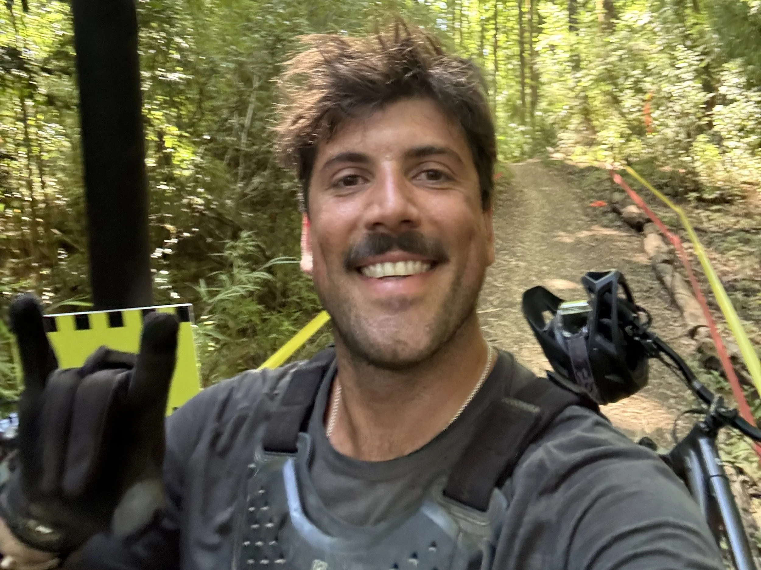 Mann tar selfie i skog med sykkel, gjør en seie med hånden, smilende, med grønt fyt og trær i bakgrunnen.