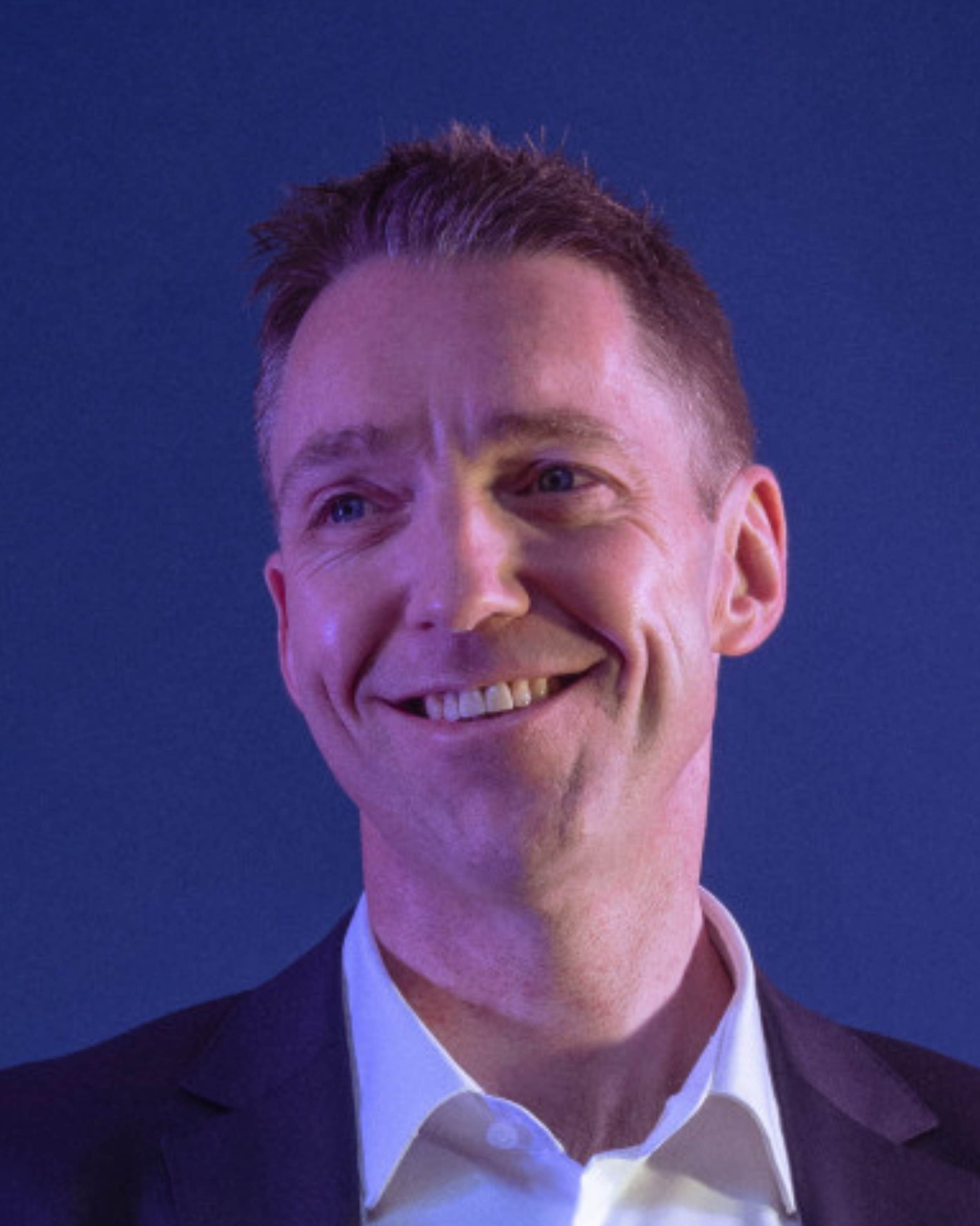 Smiling man with short hair, wearing a suit and white shirt, against a blue background.