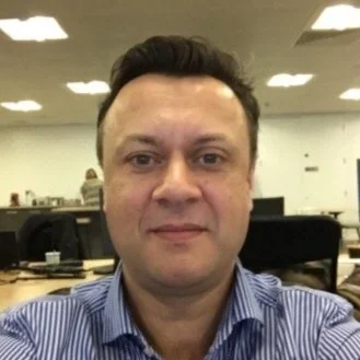 A man with short dark hair and a slight smile taking a selfie in an office environment.