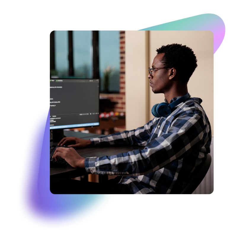 A young man with glasses and headphones around his neck working on a computer with code displayed on the screen in a modern workspace.
