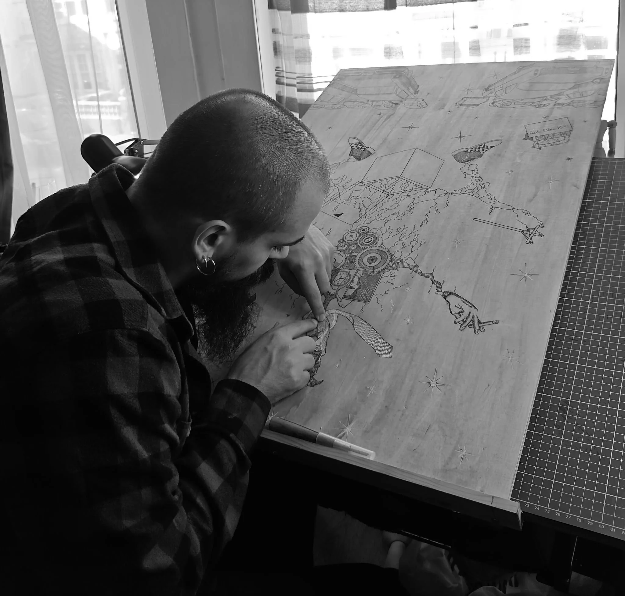 A person carving intricate designs on a wooden board at a desk in black and white.