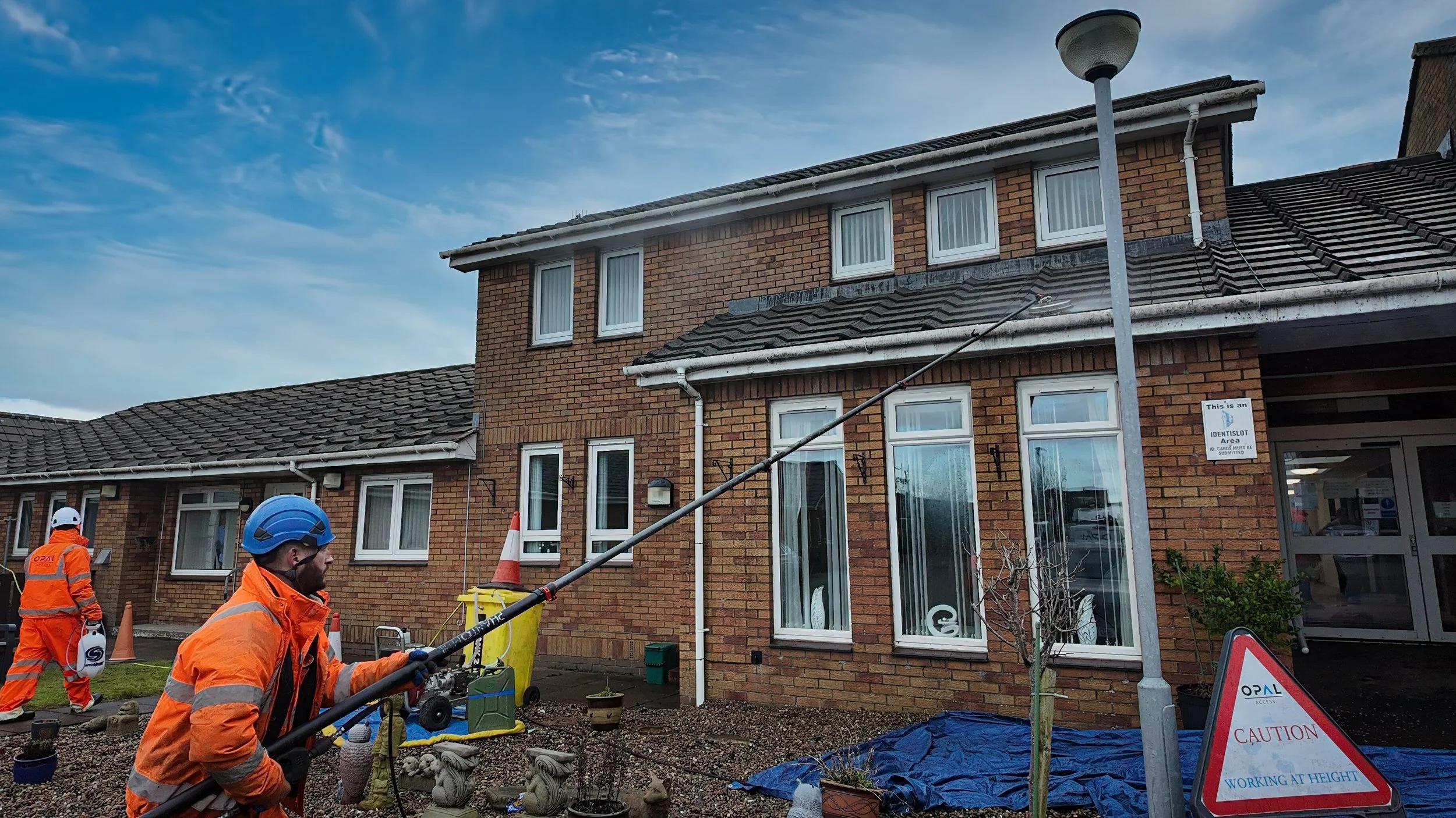 Workers in orange safety gear and helmets repairing or inspecting a brick apartment building, using an extended pole, in a parking lot with warning signs and equipment