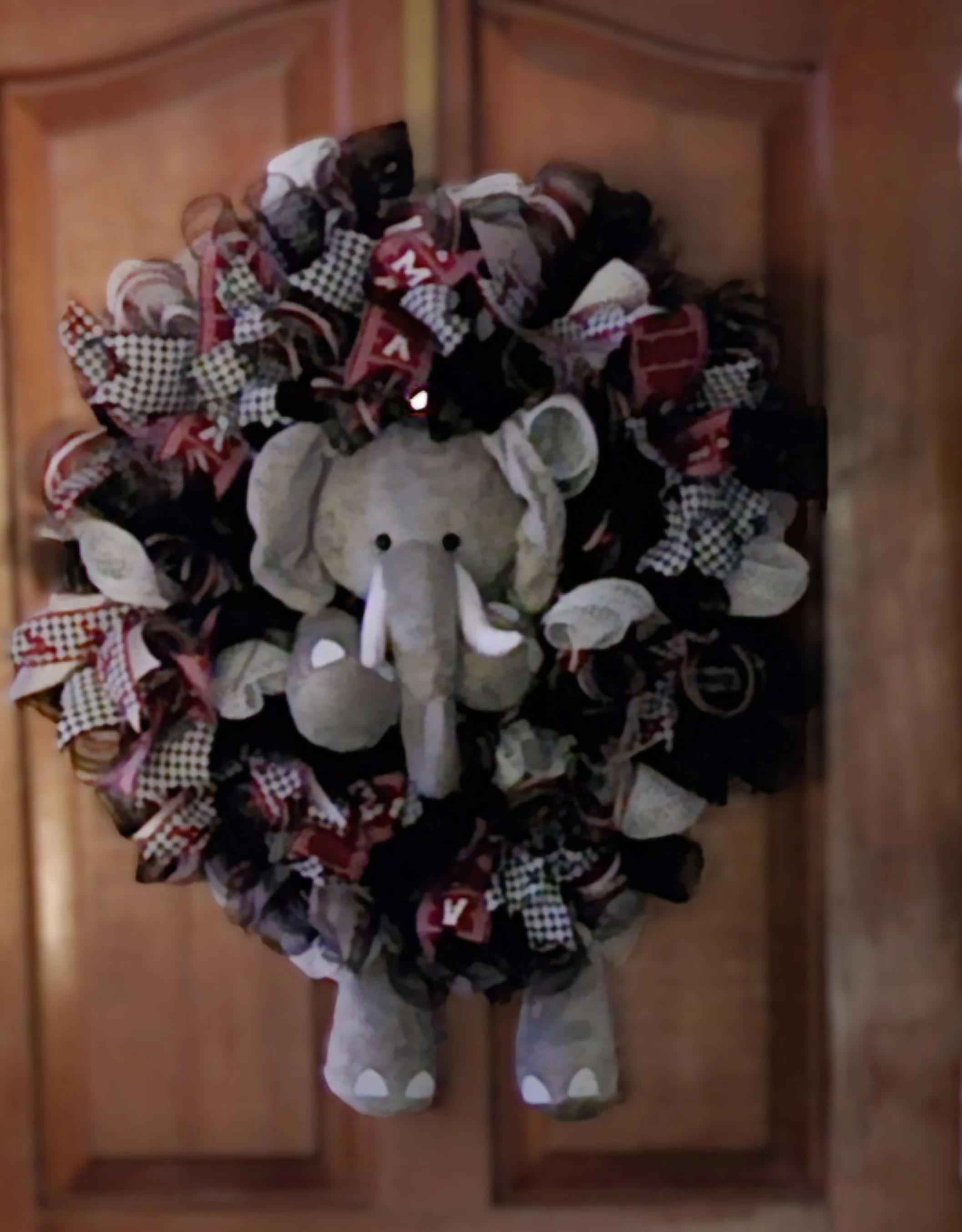 A decorative wreath with a plush elephant in the center, made of fabric ribbons in red, black, white, and checkered patterns, hanging on a wooden cabinet door.