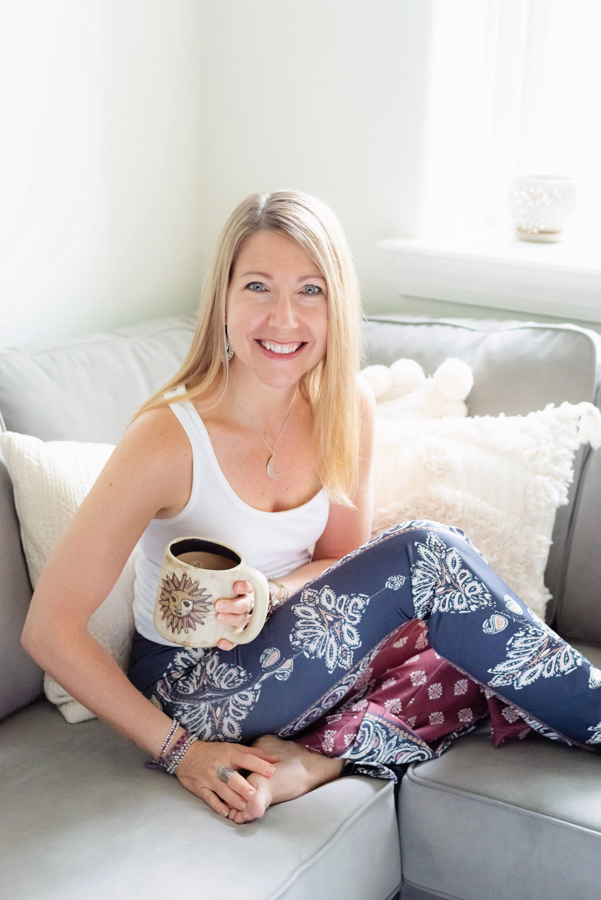 Parenting Astrologer, Tara Vogel, wearing a white tank top and patterned pants, sitting on a light-colored couch, smiling, holding a mug with a sun design, in a bright living room with white pillows and a window in the background.