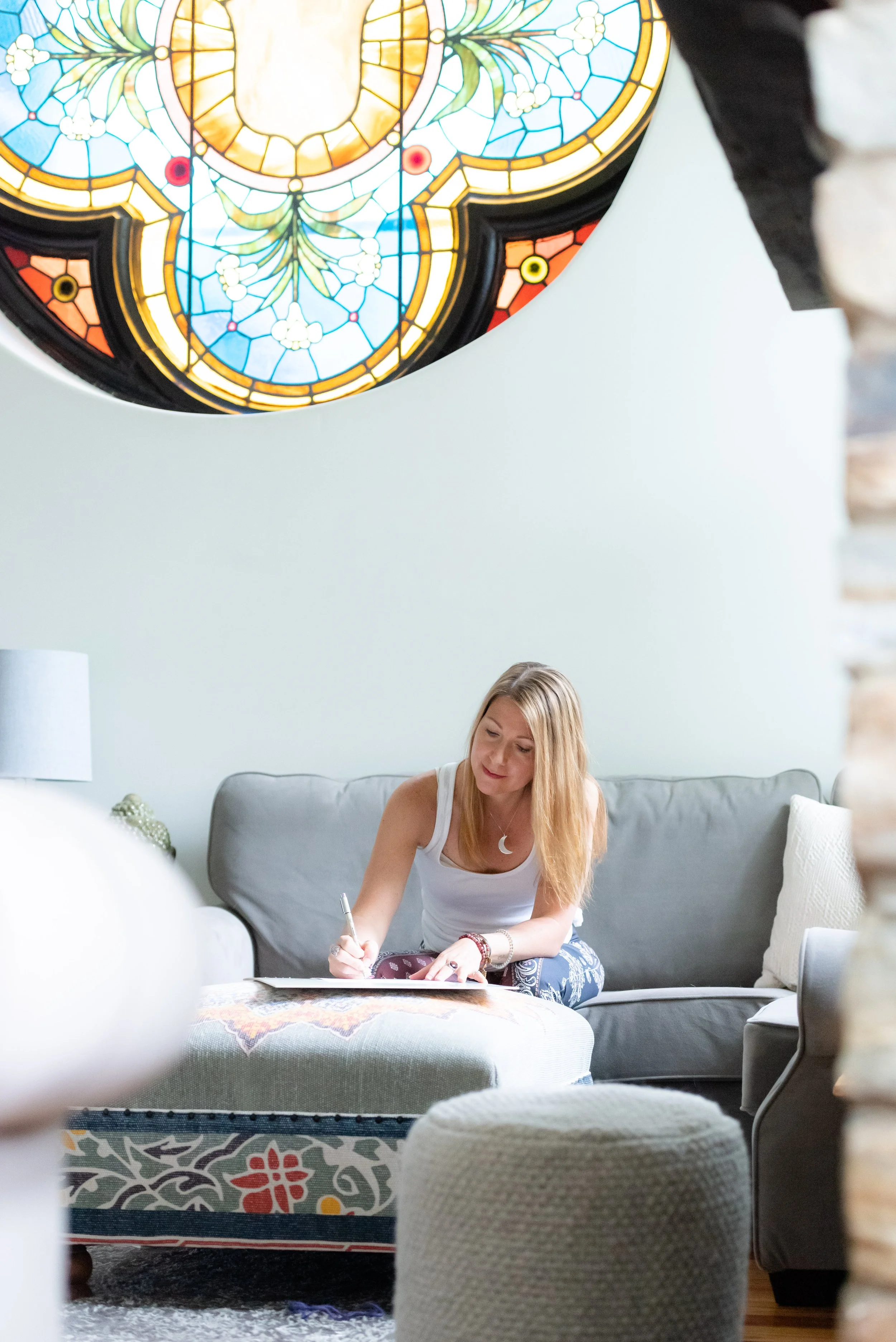 Parenting Astrologer, Tara Vogel, a woman with long blonde hair sitting on a gray couch, writing in a notebook or journal, in a well-lit living room with a stained glass lamp hanging from the ceiling.
