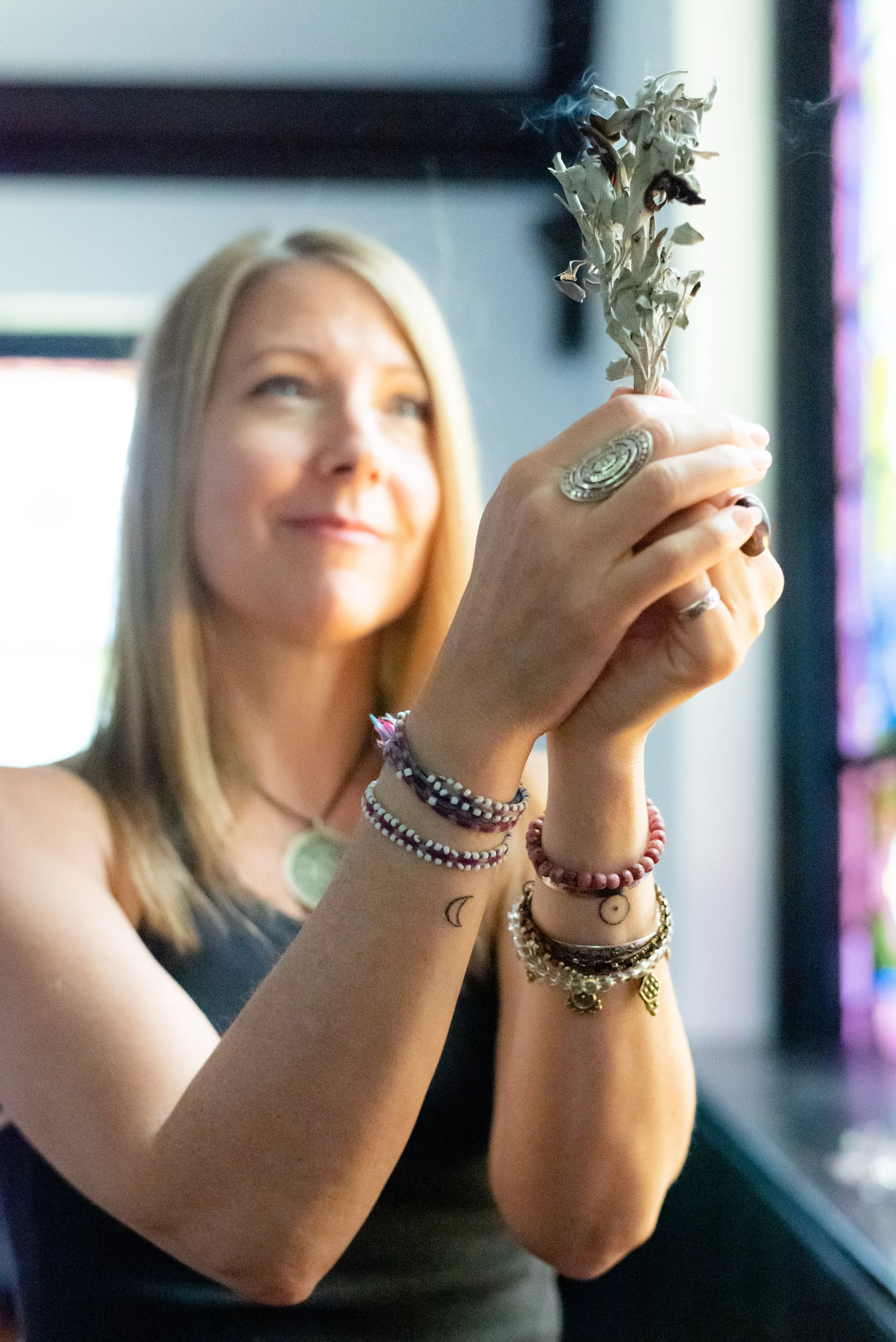 Parenting Astrologer, Tara Vogel, a woman with blonde hair holds a burning incense stick, smiling slightly, wearing multiple bracelets and rings, with tattoos on her arm, in a softly lit room.