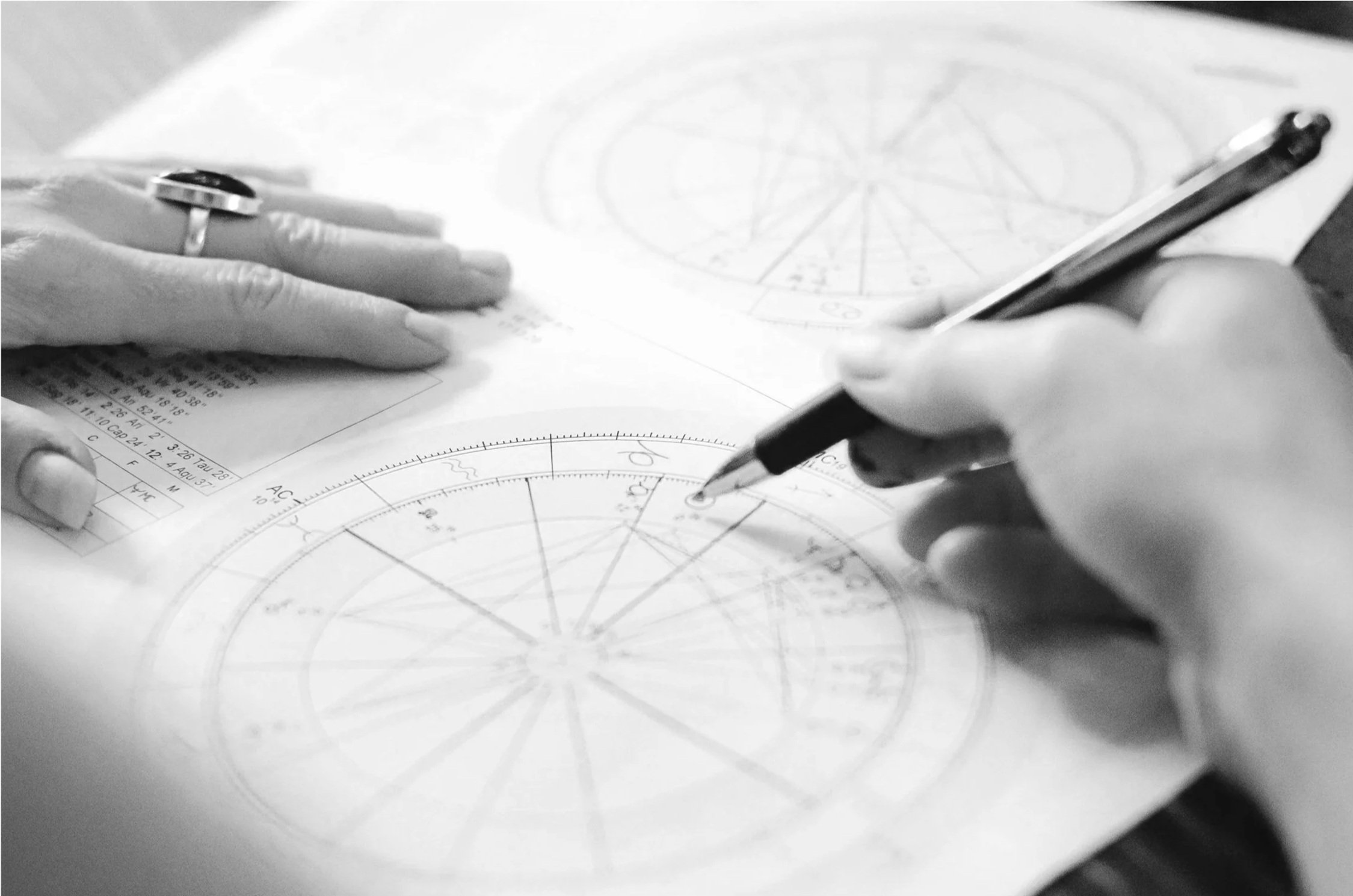 Two hands working on a detailed scientific diagram involving circles and radial lines, with one hand holding a pen and the other resting on the paper, on a desk alongside additional papers.
