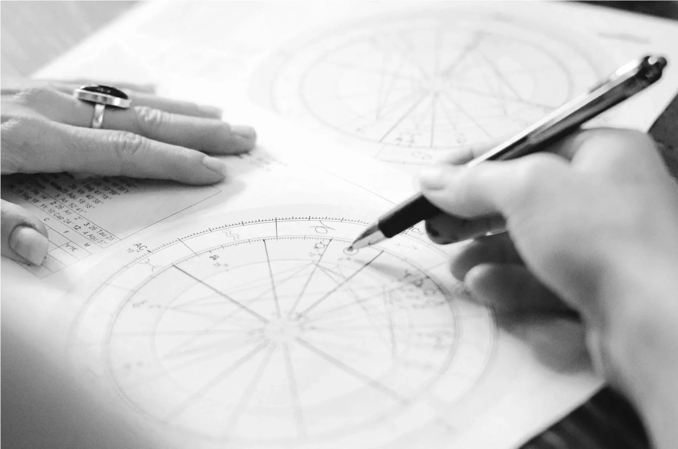 Someone is working on a detailed astrological chart or star chart, using a pen on a paper with circular diagrams and star charts, with another person's hand resting nearby.