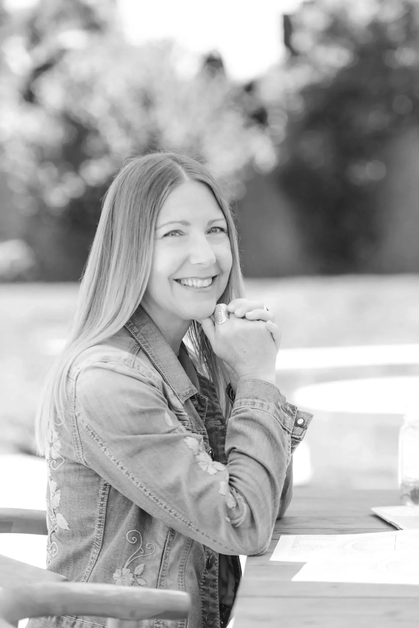 A woman smiling outdoors, seated at a table, with her hands clasped under her chin, wearing a denim jacket, and a ring on her finger.