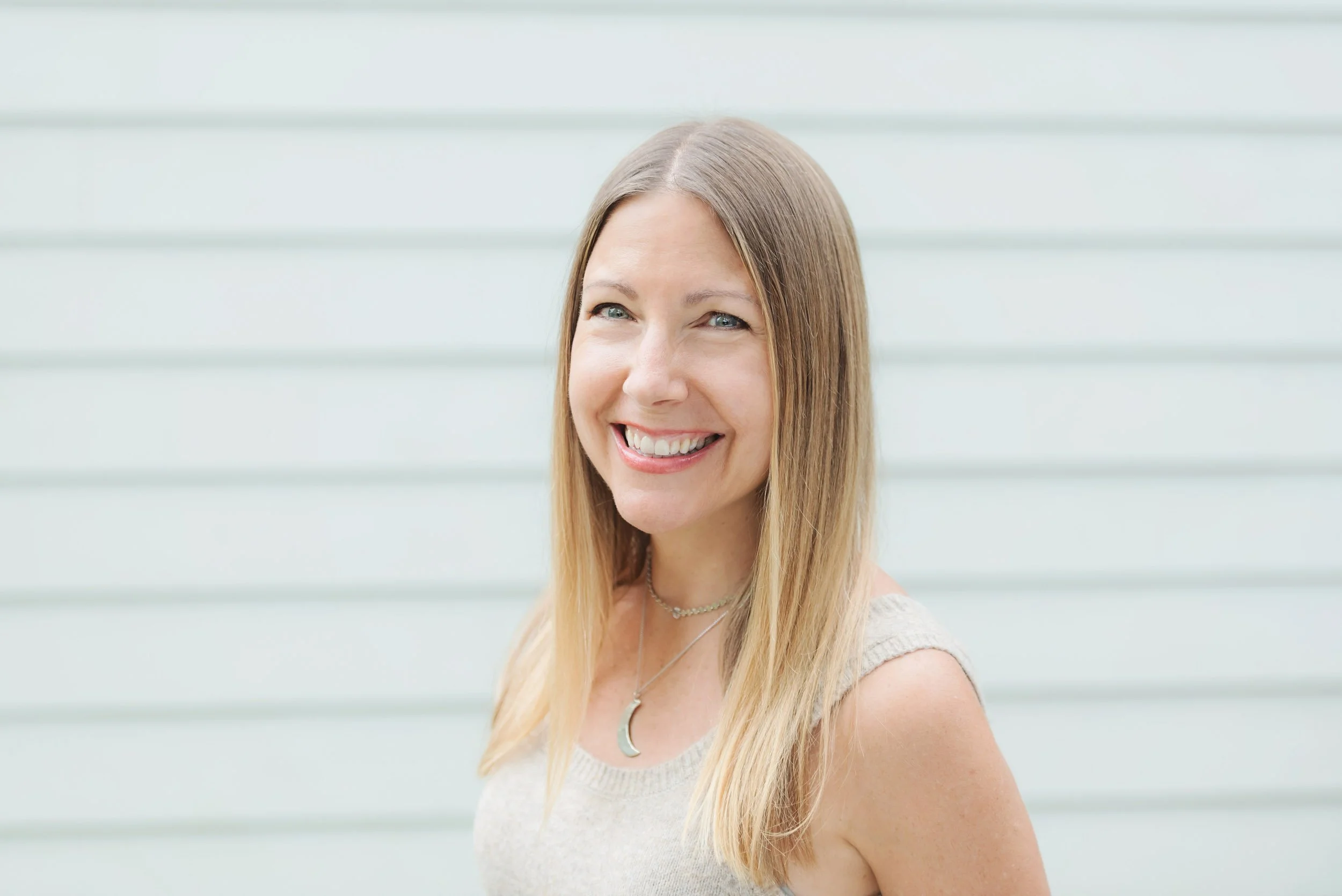 Parenting Astrologer, Tara Vogel, a woman smiling with light brown hair and blue eyes, wearing a sleeveless beige top and layered necklaces, standing in front of a white siding wall.