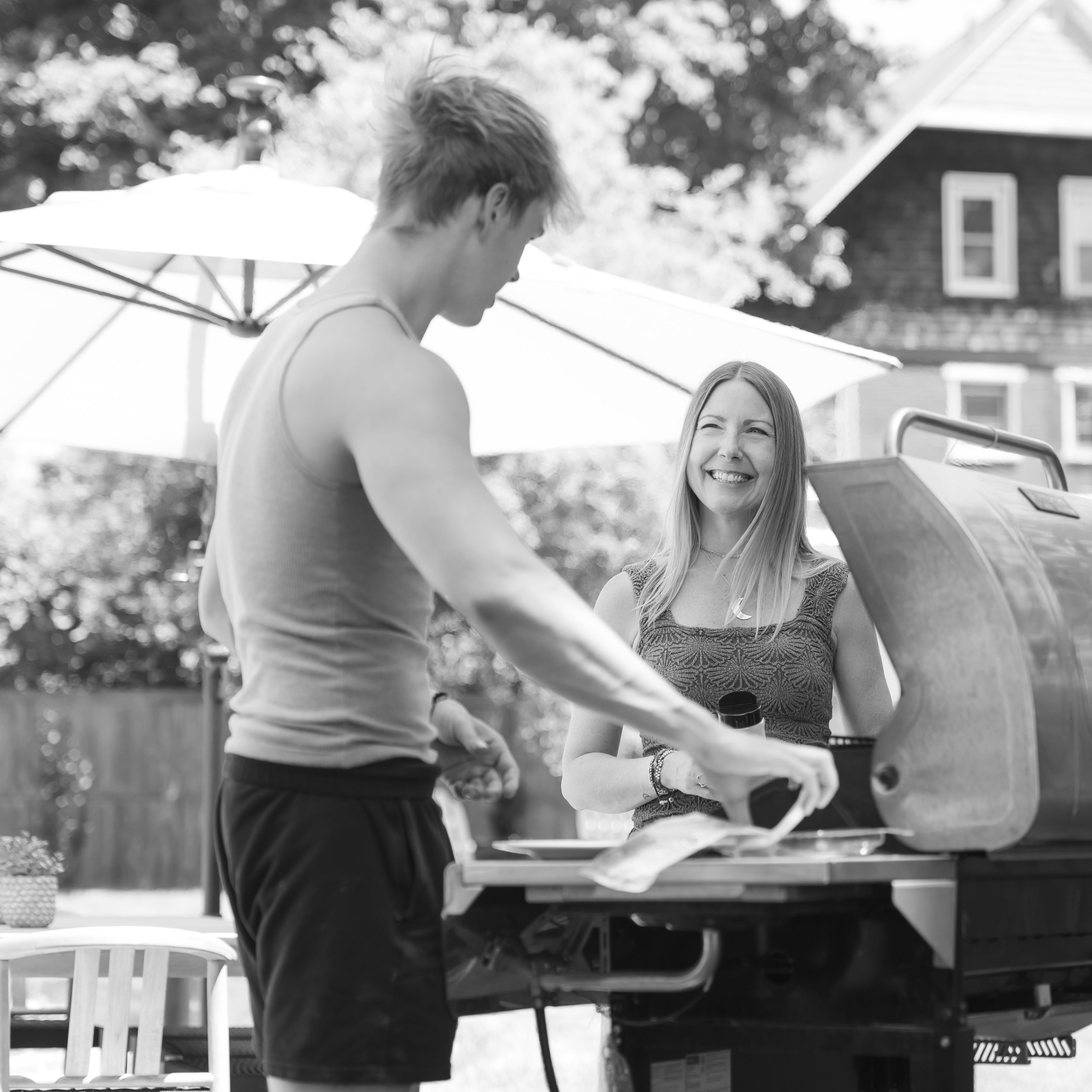 A woman smiling at a man at an outdoor gathering near a grill with an umbrella in the background.