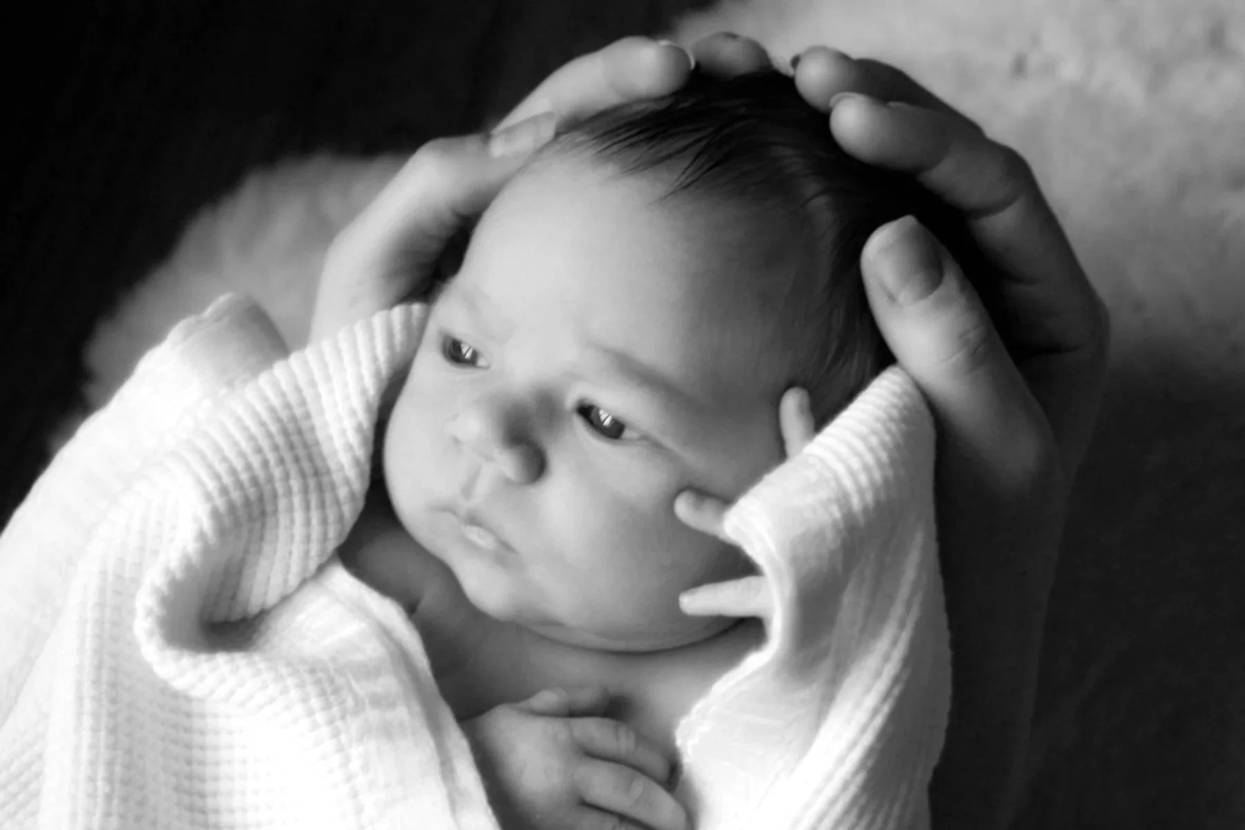 A person with adult fingers holding a baby's head, cradling it gently. The baby has a calm expression and is wearing a textured, light-colored garment. The image is in black and white.