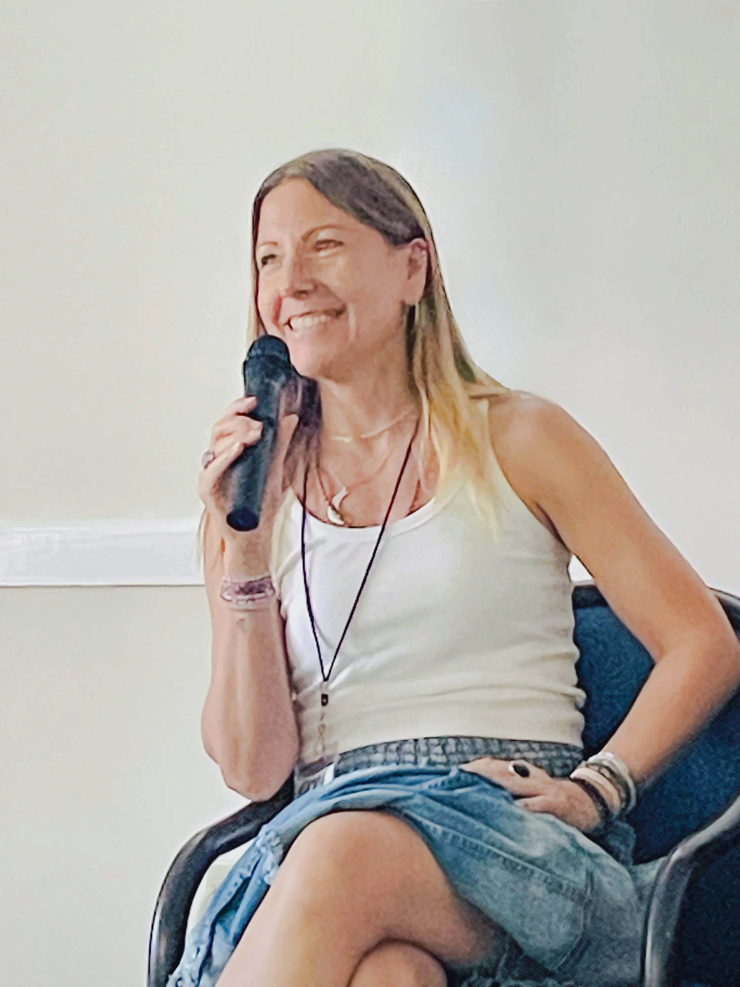 Parenting Astrologer, Tara Vogel, sitting in a chair, smiling, holding a microphone in her right hand, wearing a tank top and shorts.