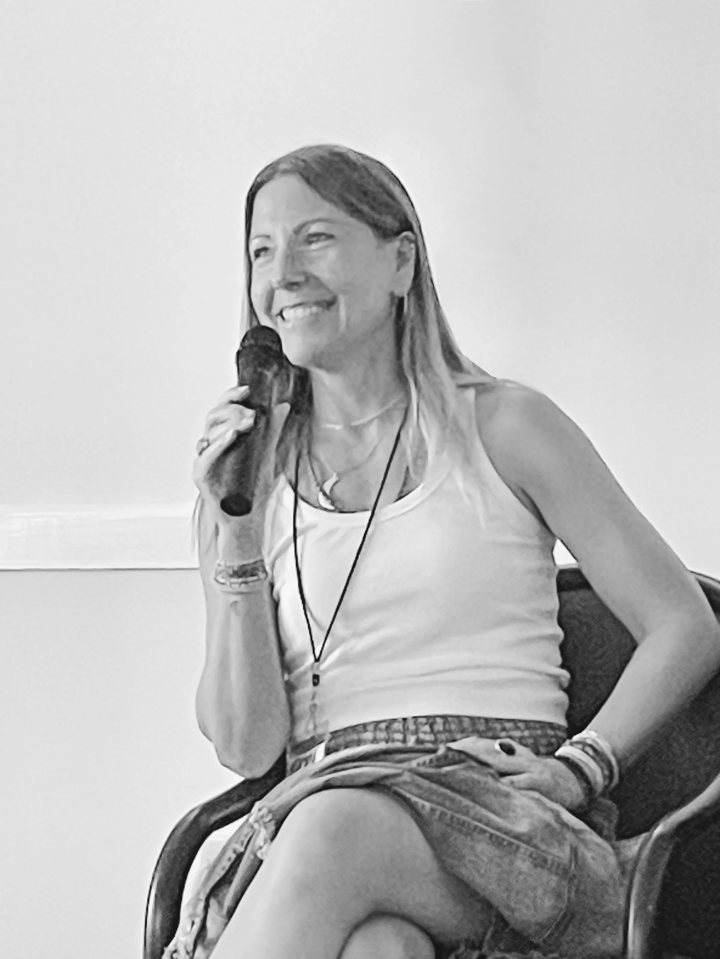 Parenting Astrologer, Tara Vogel, a woman sitting on a chair, holding a microphone, smiling, wearing a white tank top and shorts, with bracelets and necklaces.
