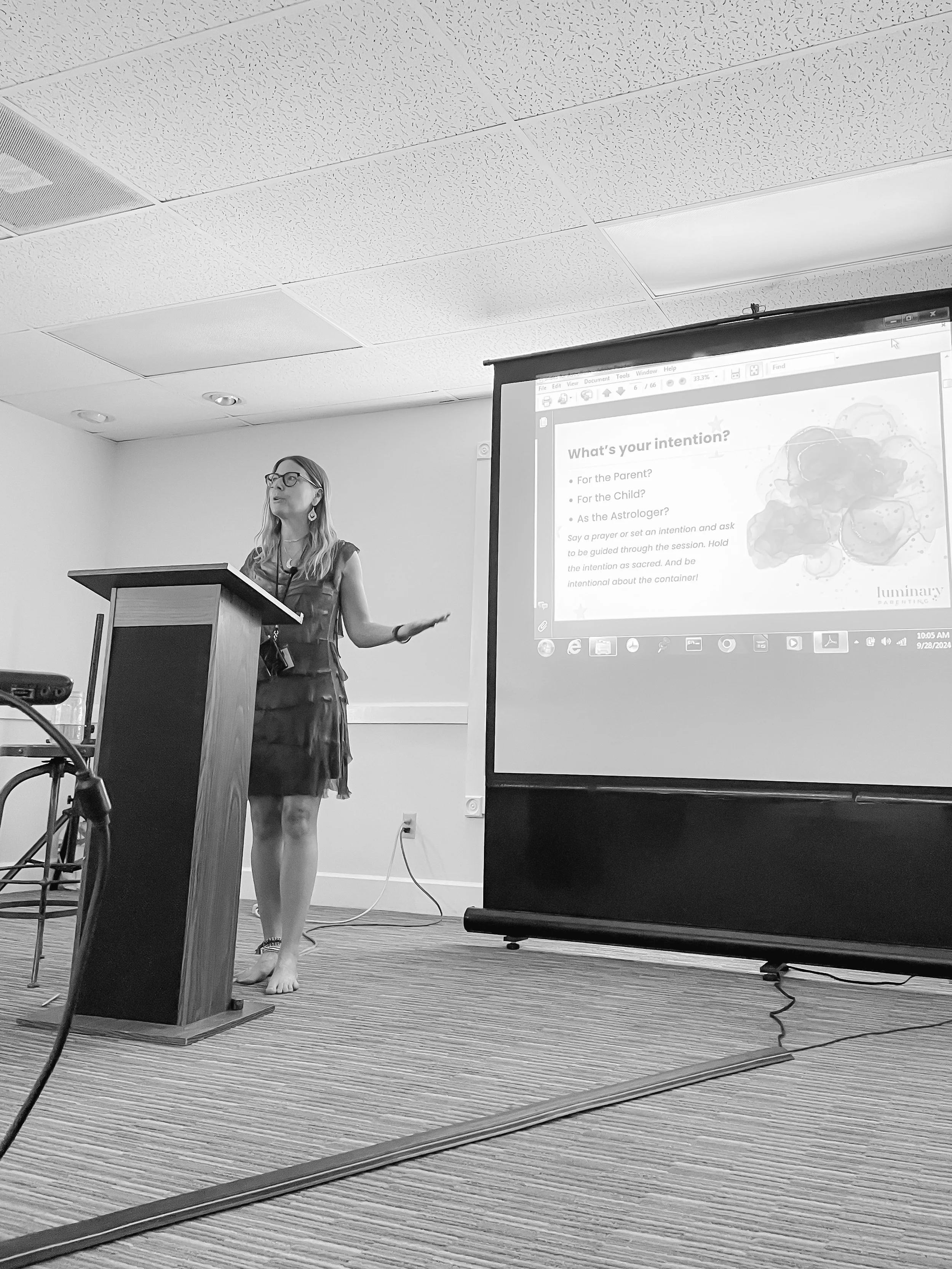 A woman in a dress and glasses is giving a presentation at a podium in a conference room. She is barefoot. There is a projector screen displaying a slide titled "What's your intention?" with bullet points and a watercolor-style image on the slide.