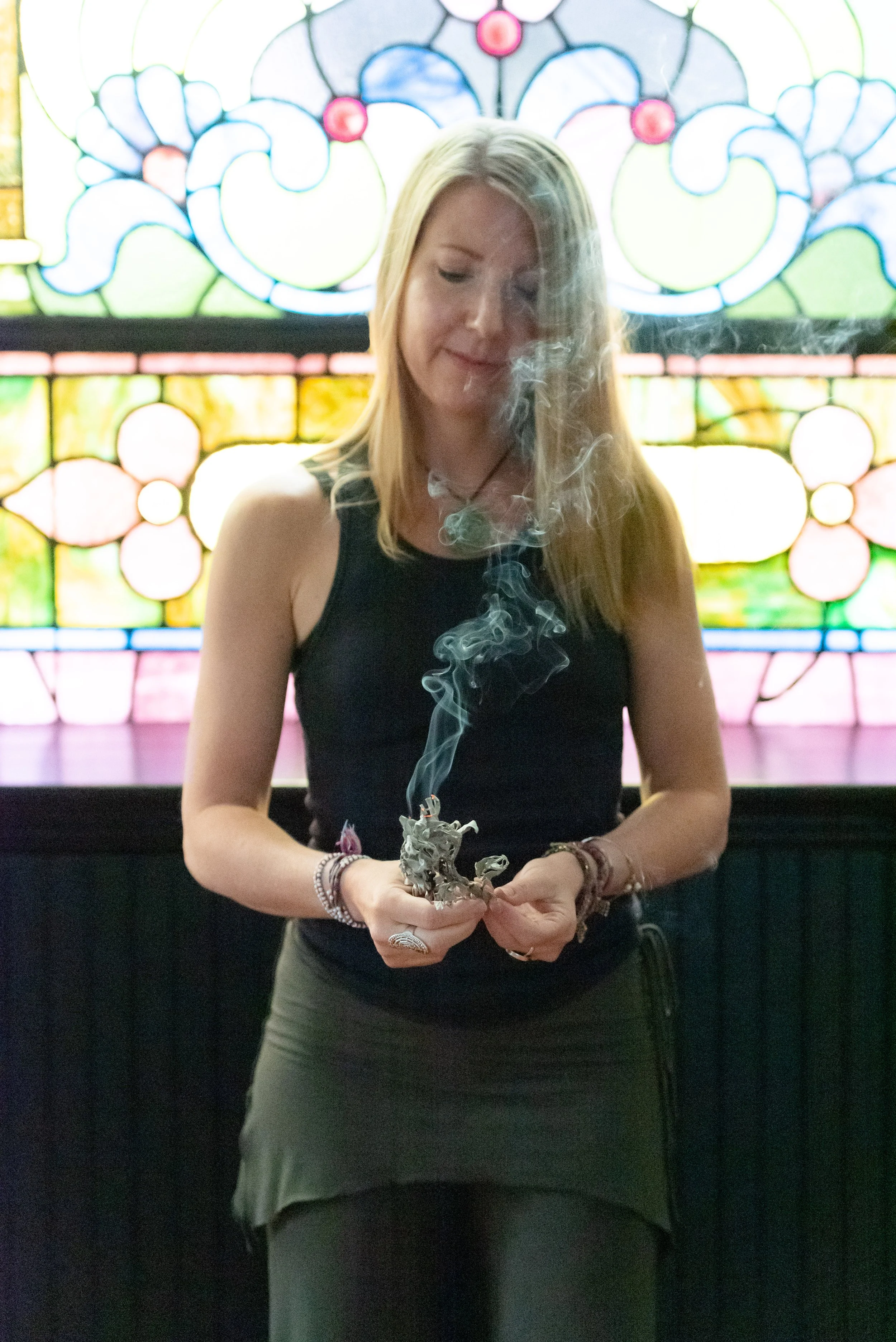 Parenting Astrologer Tara Vogel, with blonde hair, in a black sleeveless top and green skirt, stands in front of a stained-glass window, holding a smoldering sage bundle, with smoke rising from it.