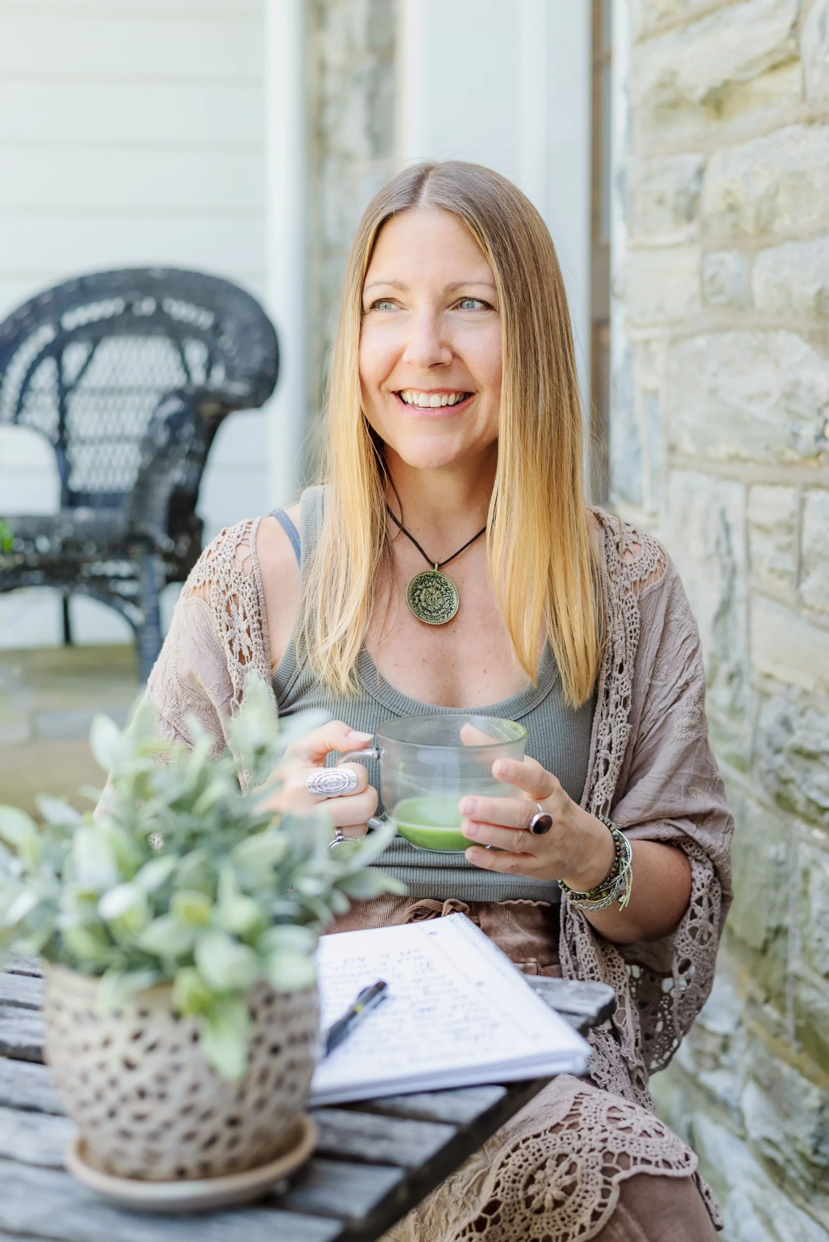 Astrologer, Tara Vogel, outdoors on a wooden table, holding a green mug. She wears a gray top, lace cardigan, a circular pendant necklace, and bracelets. A notebook and pen rest on the table, with a potted plant, wicker chairs and a stone wall.