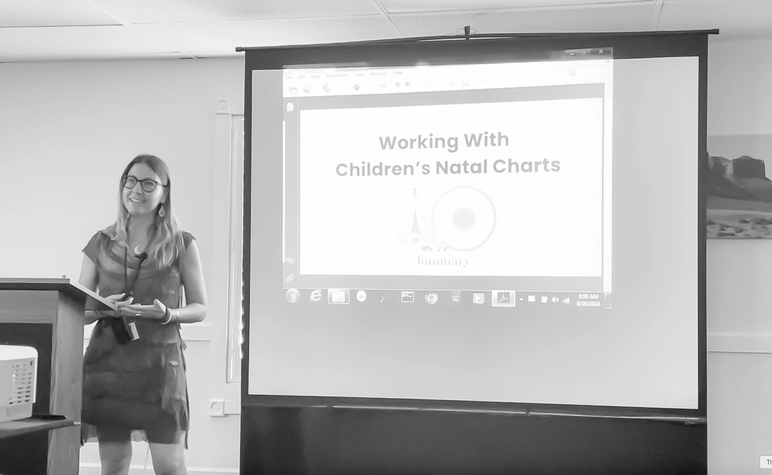 Parenting Astrologer, Tara Vogel, with glasses and long hair, standing next to a podium in a classroom or conference room, holding a booklet. Behind her is a projection screen displaying a presentation titled 'Working With Children's Natal Charts'.