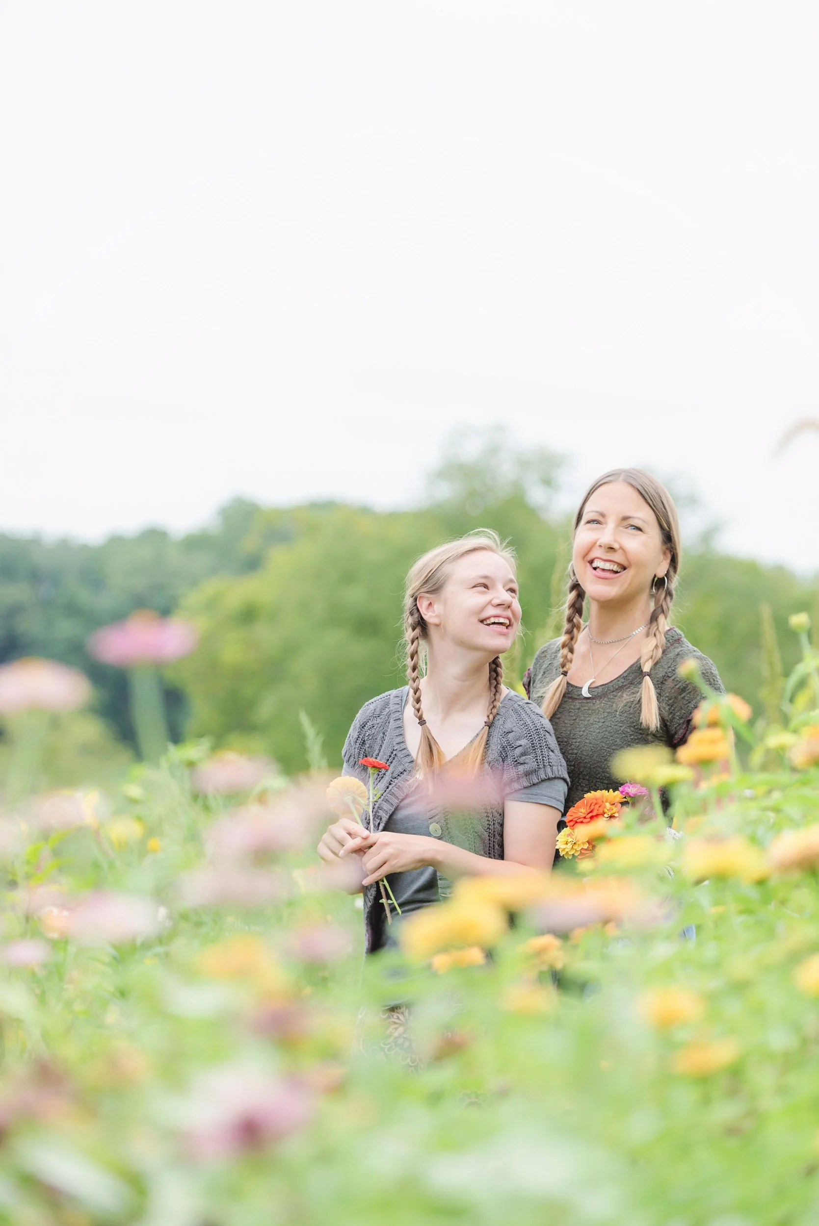 A woman and girl smiling and holding flowers in a garden filled with colorful flowers, with greenery and trees in the background.