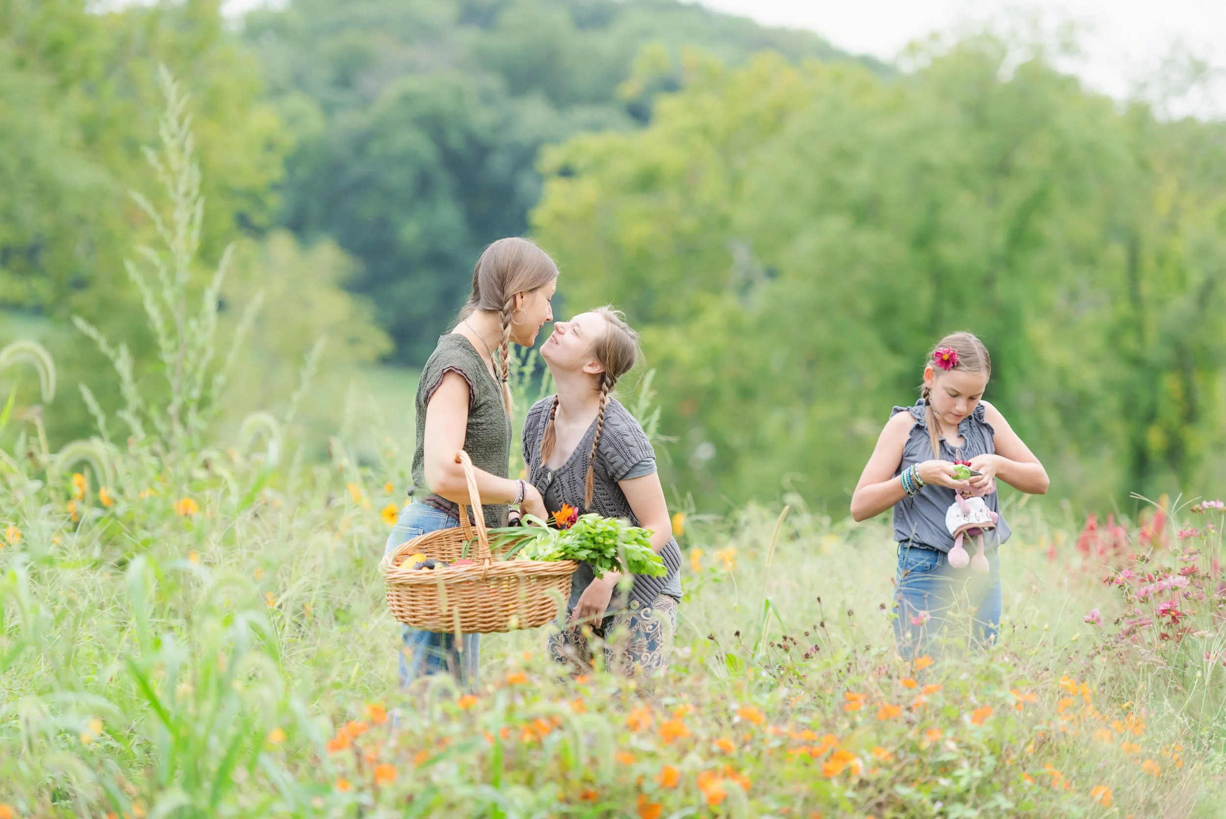 Three girls in a field of wildflowers, with two of them facing and close to each other, smiling and engaging in conversation, while the third girl on the right looks at an object in her hands, surrounded by green trees in the background.