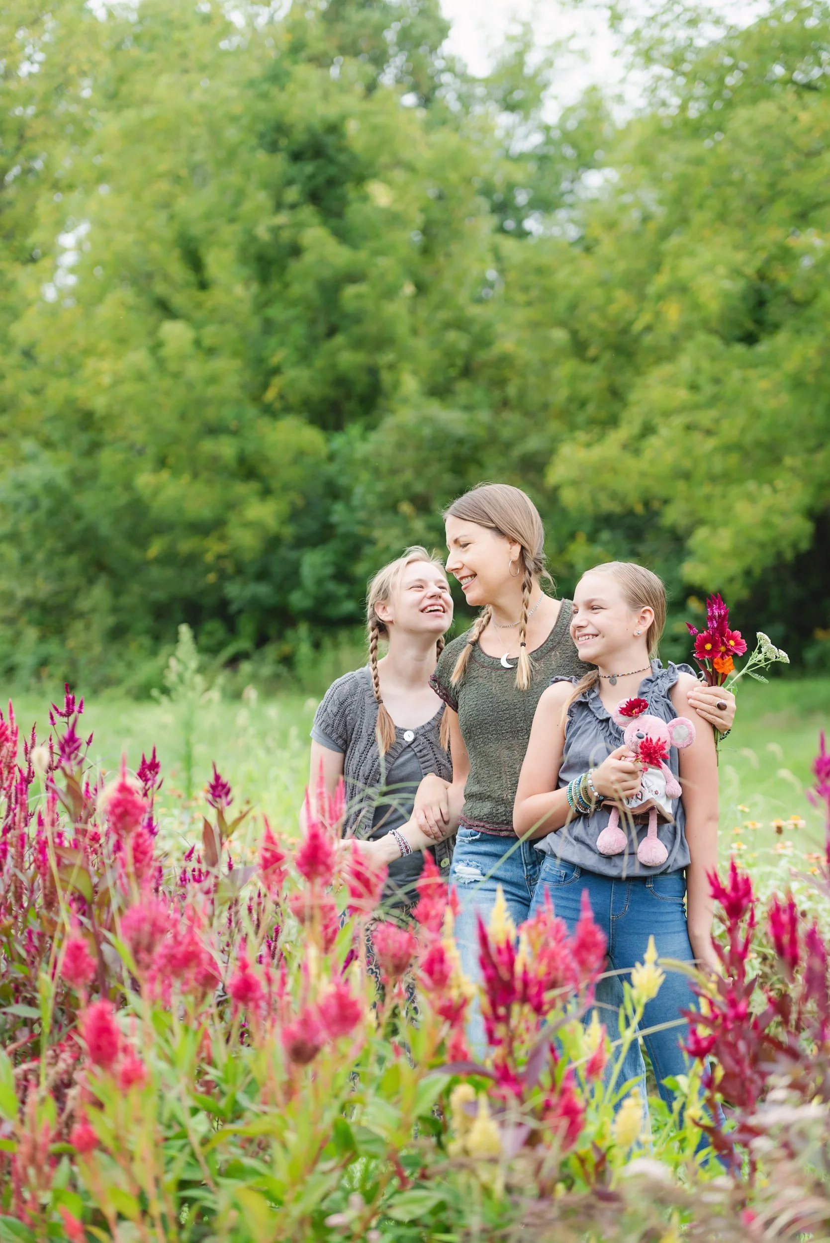 A woman and two young girls stand in a field of vibrant pink flowers, smiling and laughing together, with green trees in the background.