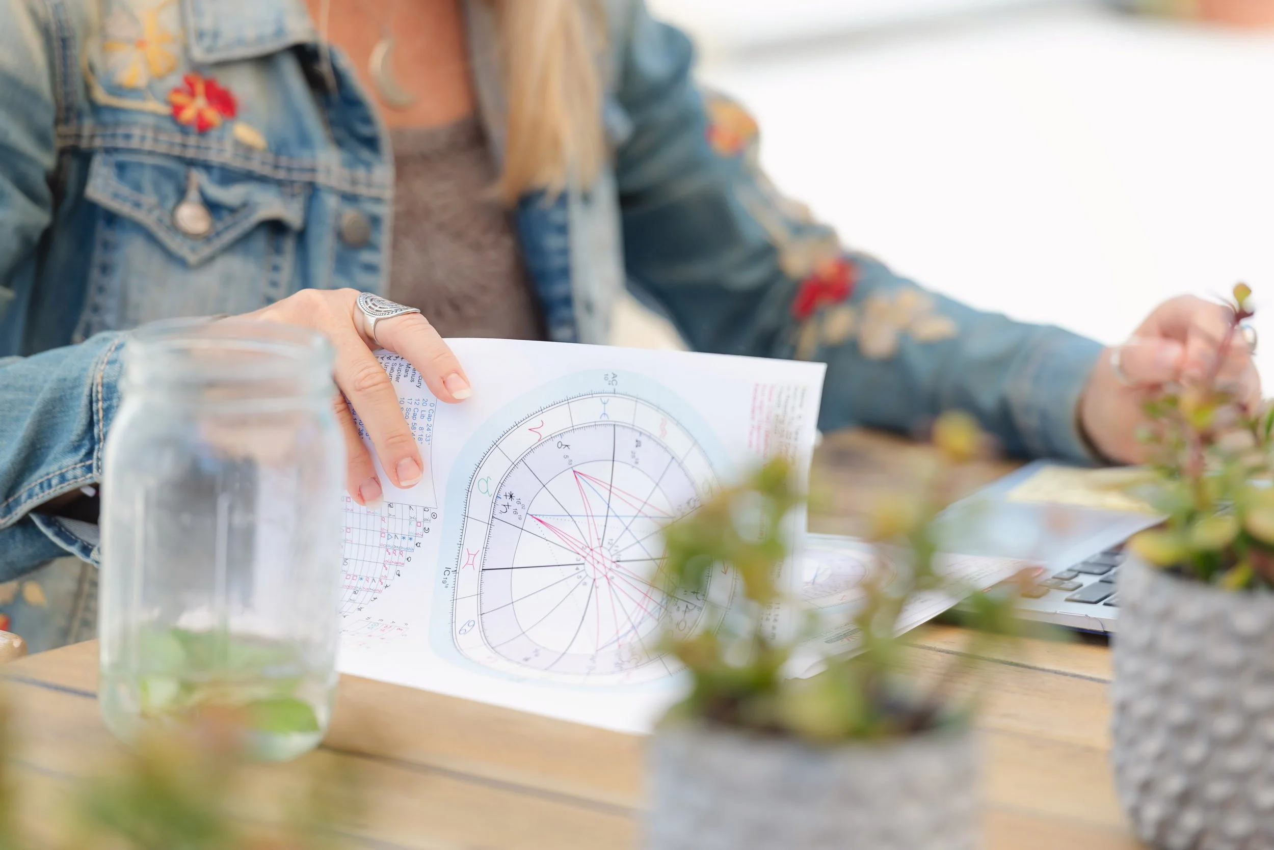 A woman sitting at a table holding an astrology chart or horoscope paper, with a glass jar and potted plants on the table, and part of a laptop visible in the background.