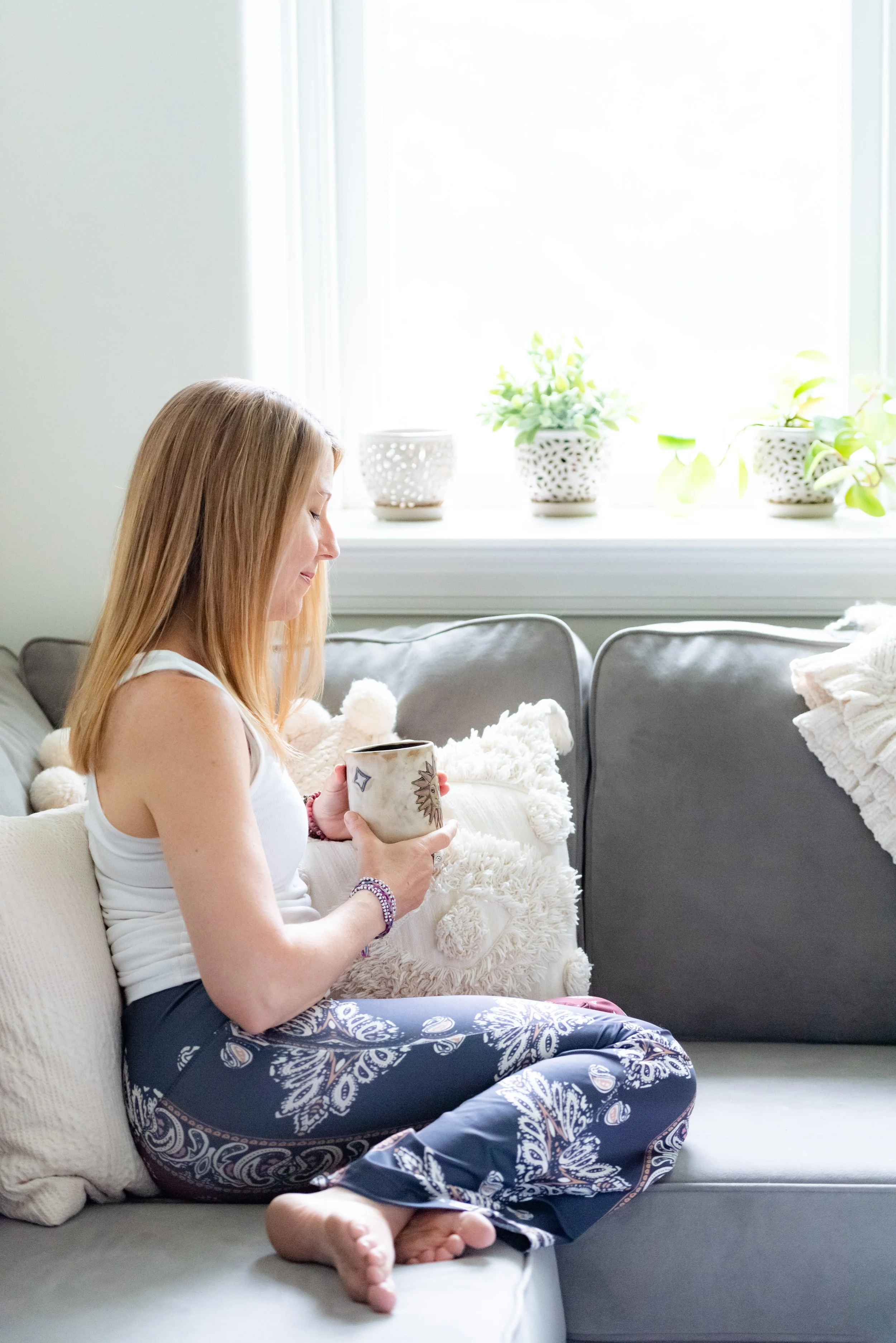 Parenting Astrologer, Tara Vogel, a woman with long blonde hair, wearing a white tank top and patterned blue pajama pants, sitting cross-legged on a gray sofa, holding a mug, with white pillows and plants on the windowsill behind her.