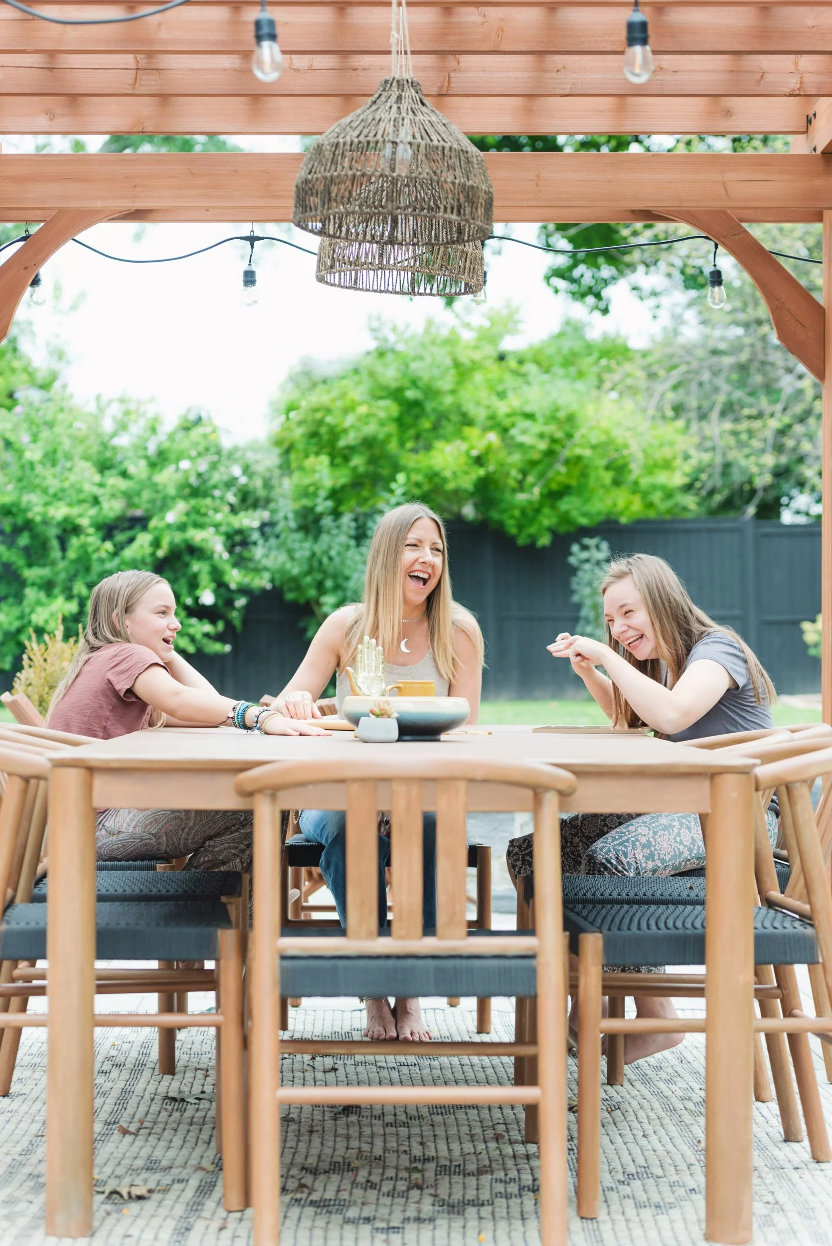 Parenting Astrologer, Tara Vogel, with your girls, sitting at a wooden outdoor table under a pergola, laughing and enjoying each other's company. The background shows greenery and a black fence.