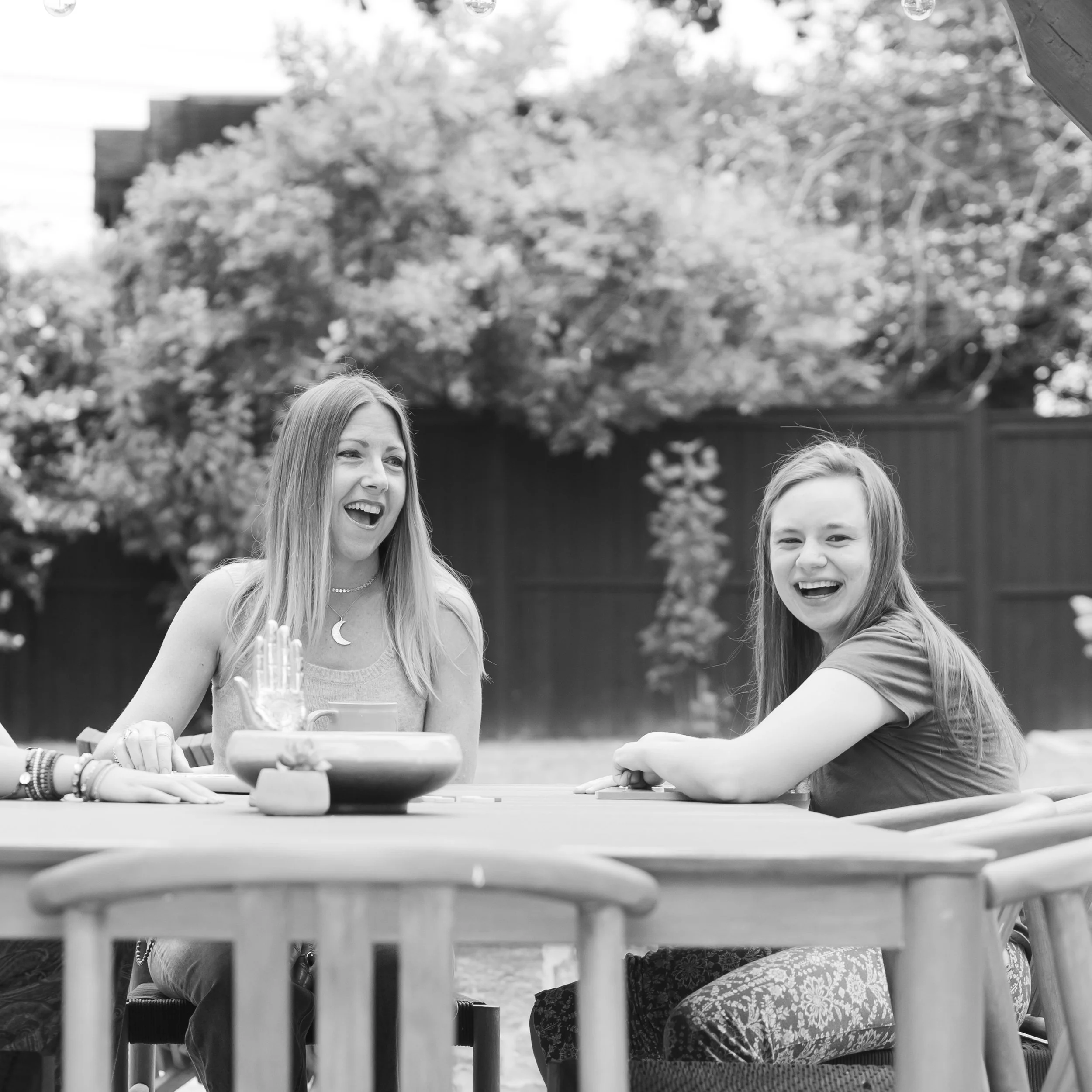 Two women sitting at a table outdoors, smiling and laughing during a social gathering.