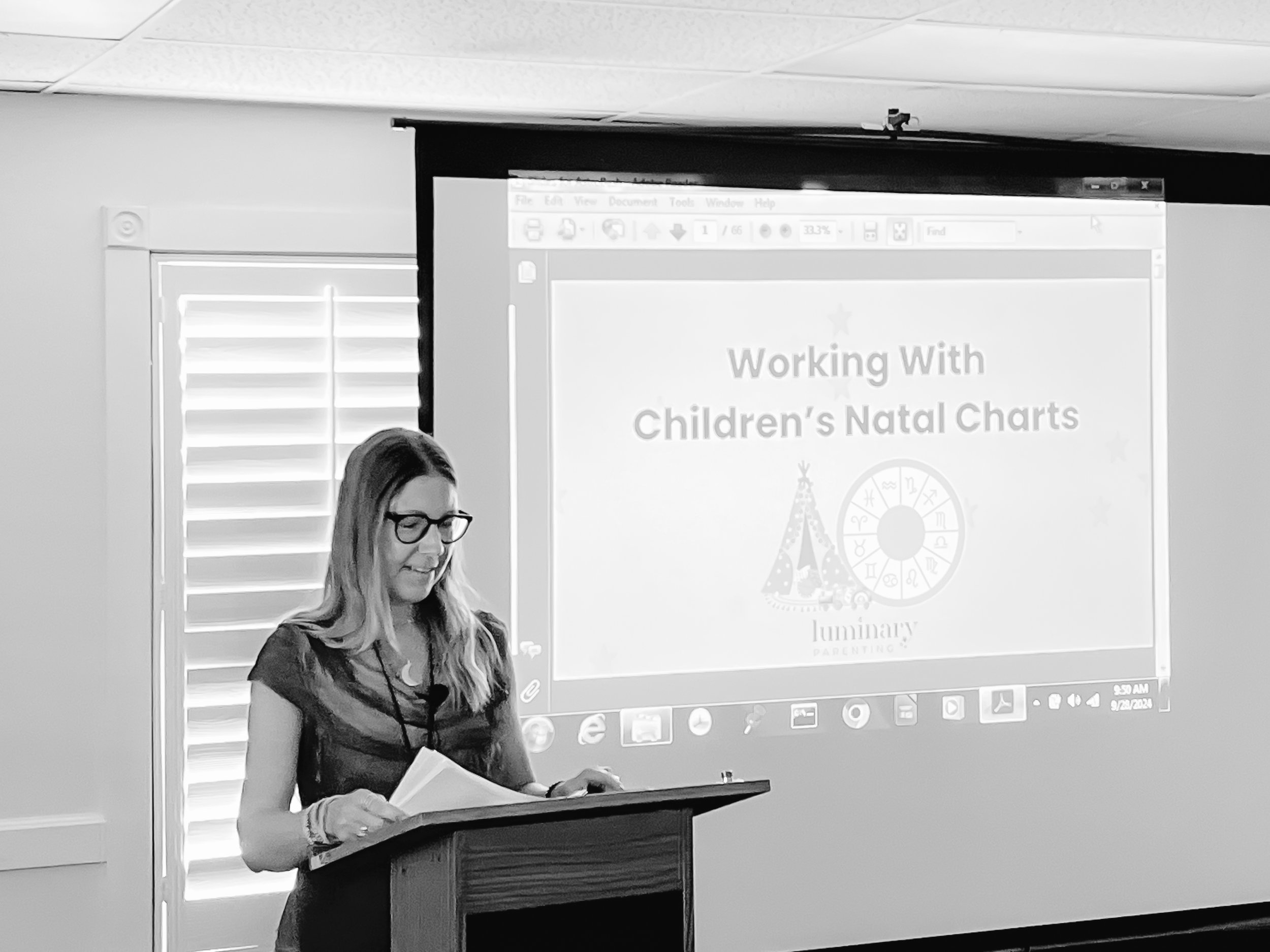 Parenting Astrologer, Tara Vogel, a woman with long hair, glasses, and a lanyard, standing at a podium. Behind her is a projection screen with a presentation titled 'Working With Children's Natal Charts' and a logo with a teepee and zodiac wheel.