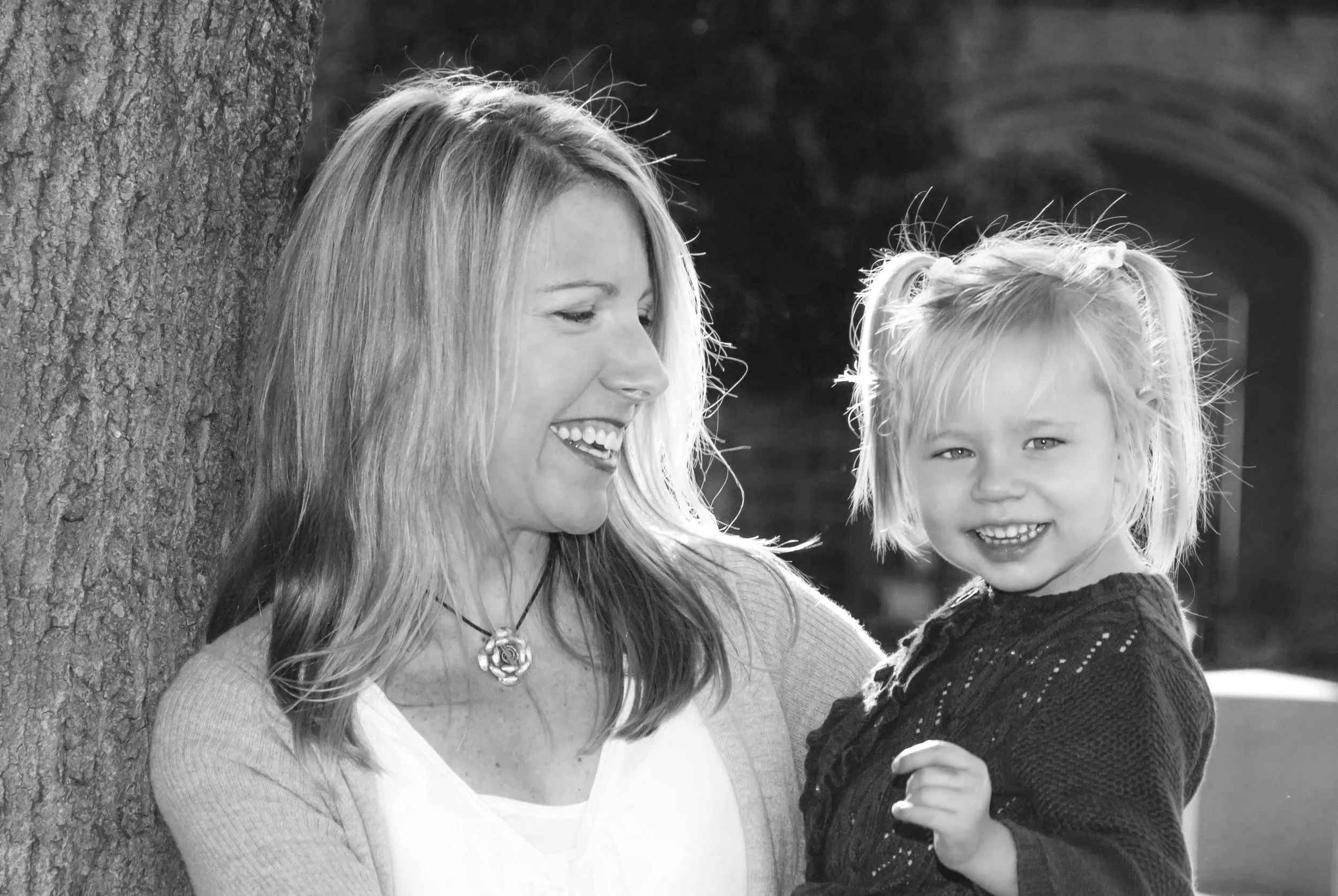 A woman with shoulder-length hair and a young girl with twin ponytails head outside, smiling and looking at each other.