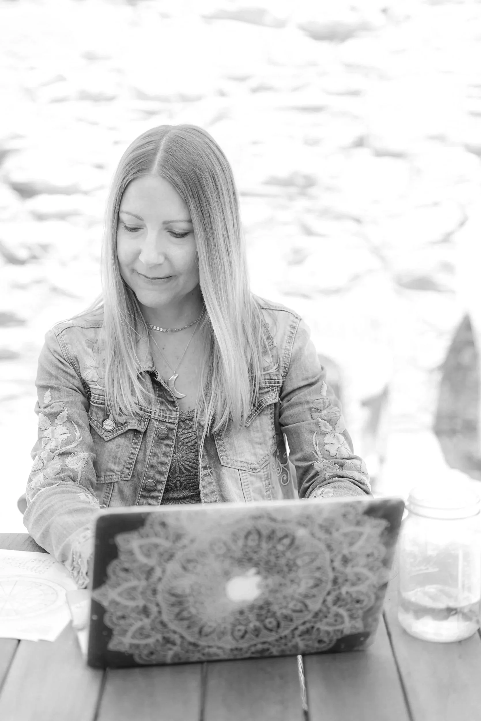 A woman with long hair sitting at a table using a laptop with a decorative cover, outdoors near a body of water.