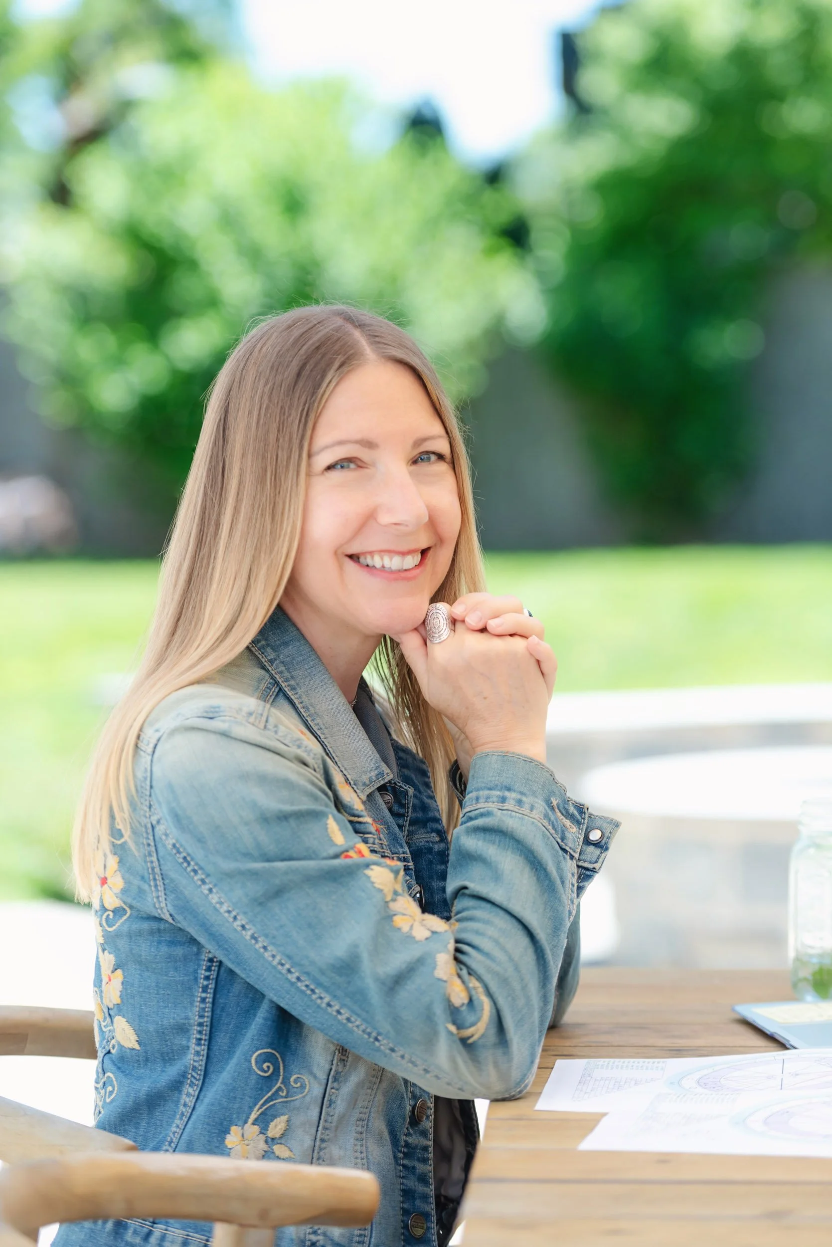 Parenting Astrologer, Tara Vogel, smiling with long blonde hair, wearing a denim jacket with floral embroidery, sitting at an outdoor table with papers, in a green garden.